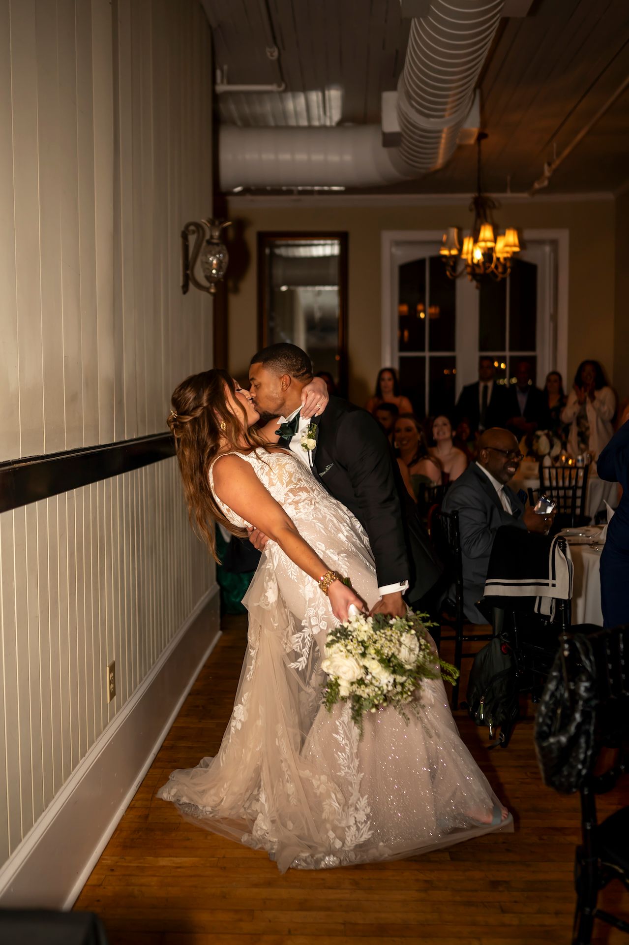 Couple shares romantic kiss during wedding reception in dimly lit venue with white decor and hardwood floors.