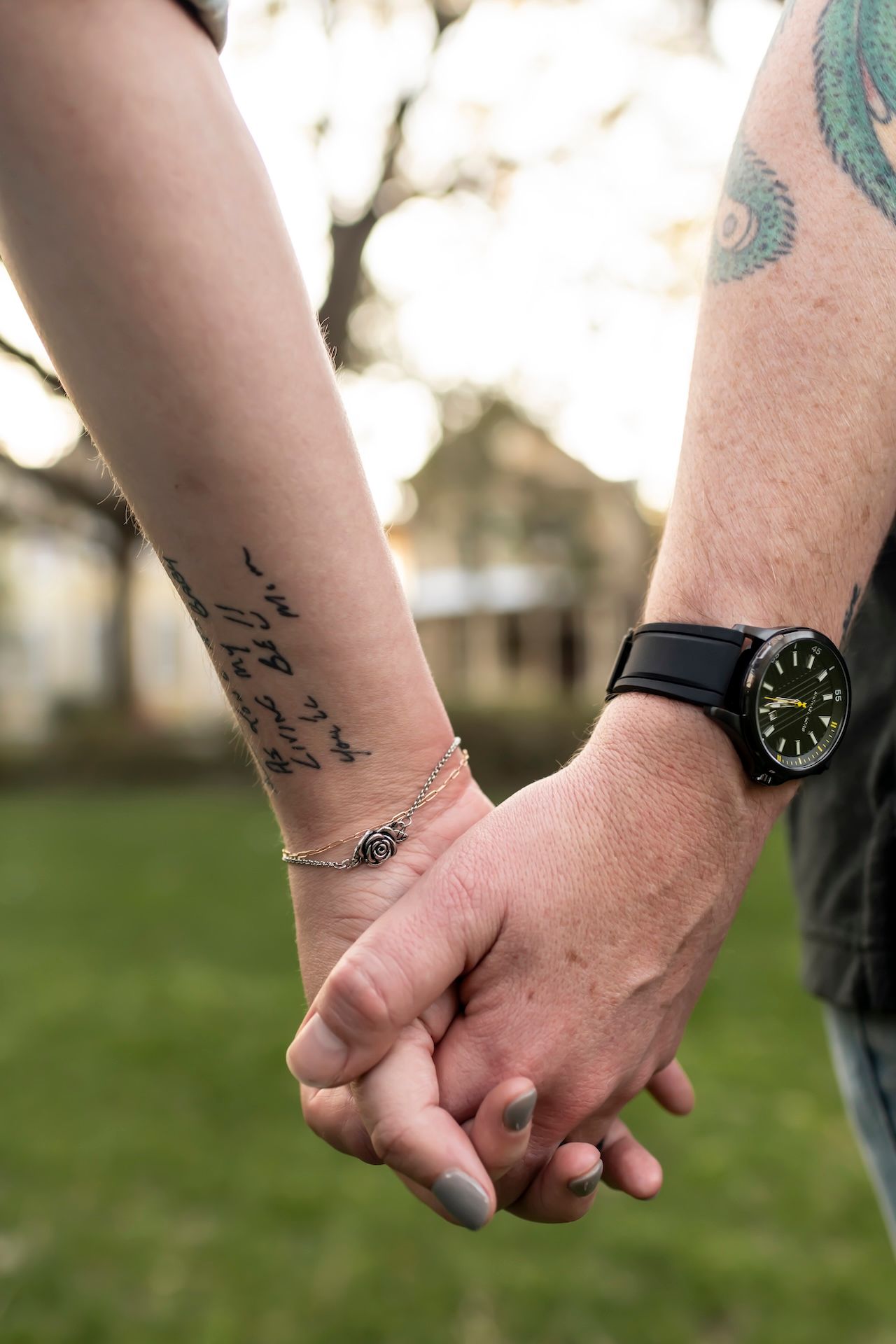 Close-up of two people holding hands outdoors, with a small tattoo and wristwatch visible.