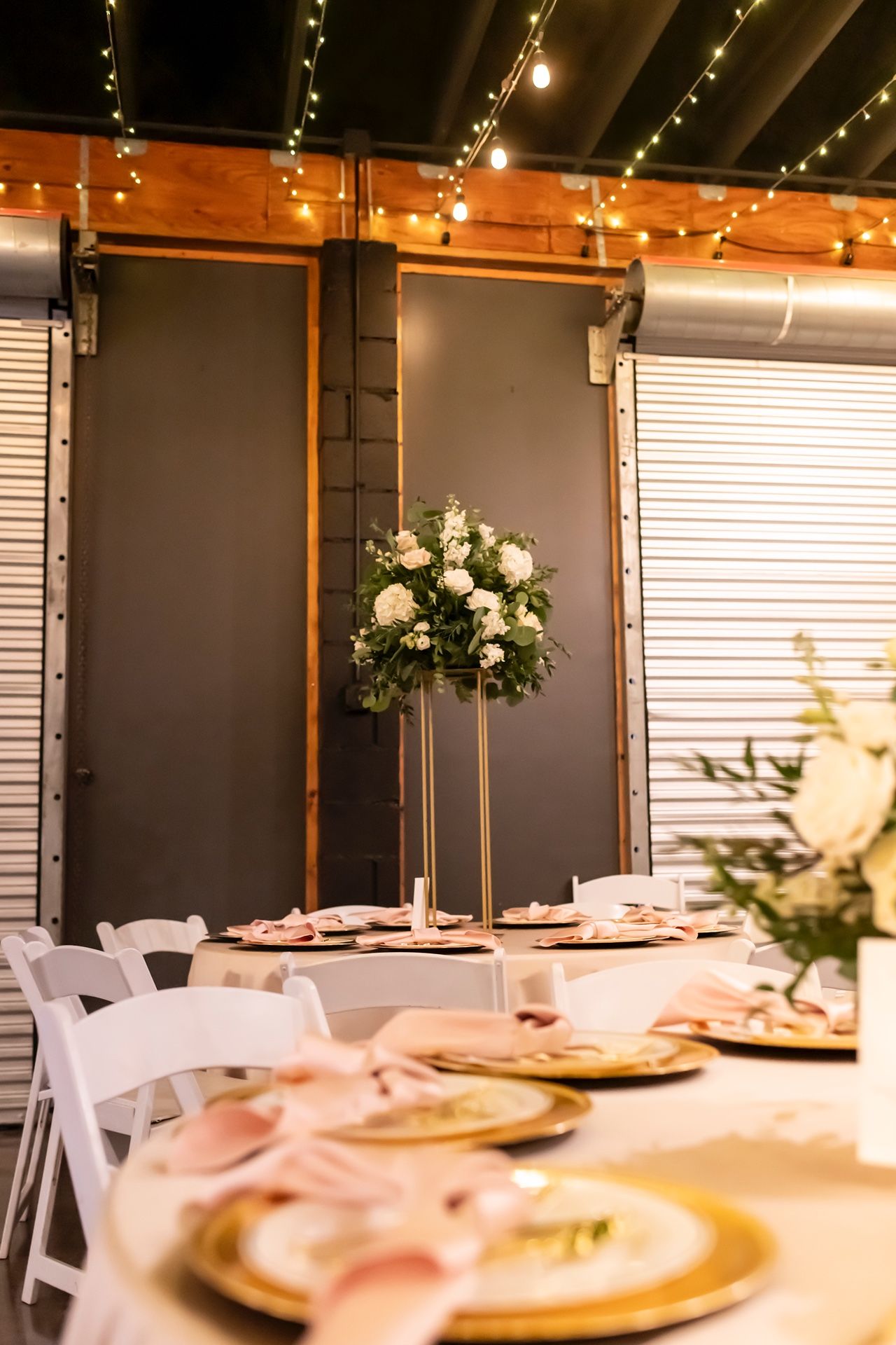 Elegant wedding reception table with gold charger plates and tall white floral centerpiece under string lights.