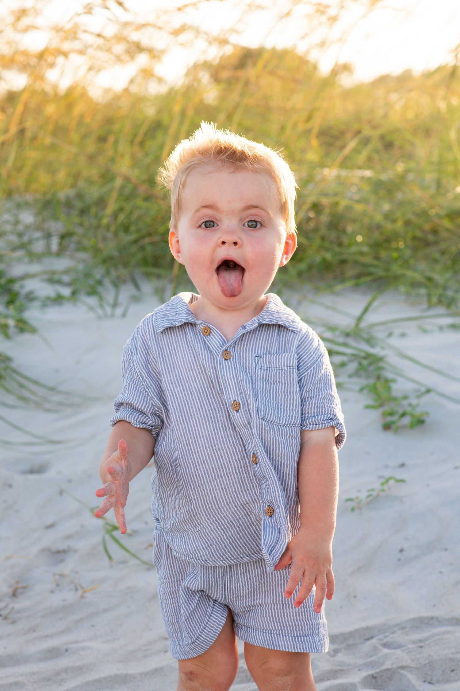 Young child wearing blue striped outfit makes playful expression while standing on sandy beach at sunset.