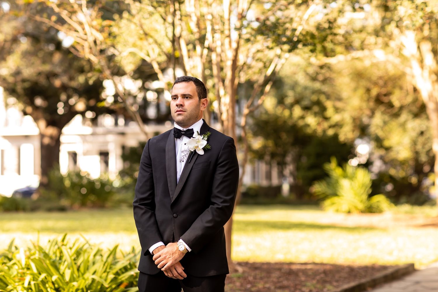 A groom in a black tuxedo and bowtie stands in front of trees during a golden sunset at an outdoor wedding.