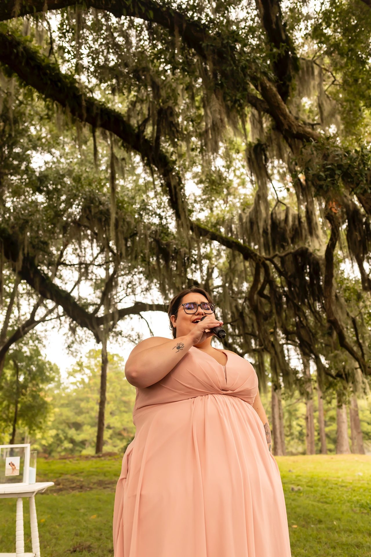 A musician in a flowing peach dress performs outdoors under moss-draped oak trees at a garden event.