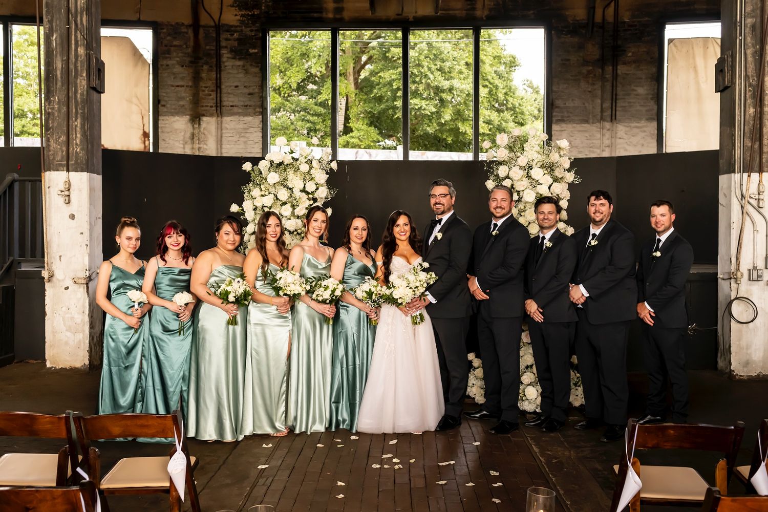 Wedding party stands in rustic venue with bridesmaids in aqua dresses and groomsmen in black suits under floral arch.