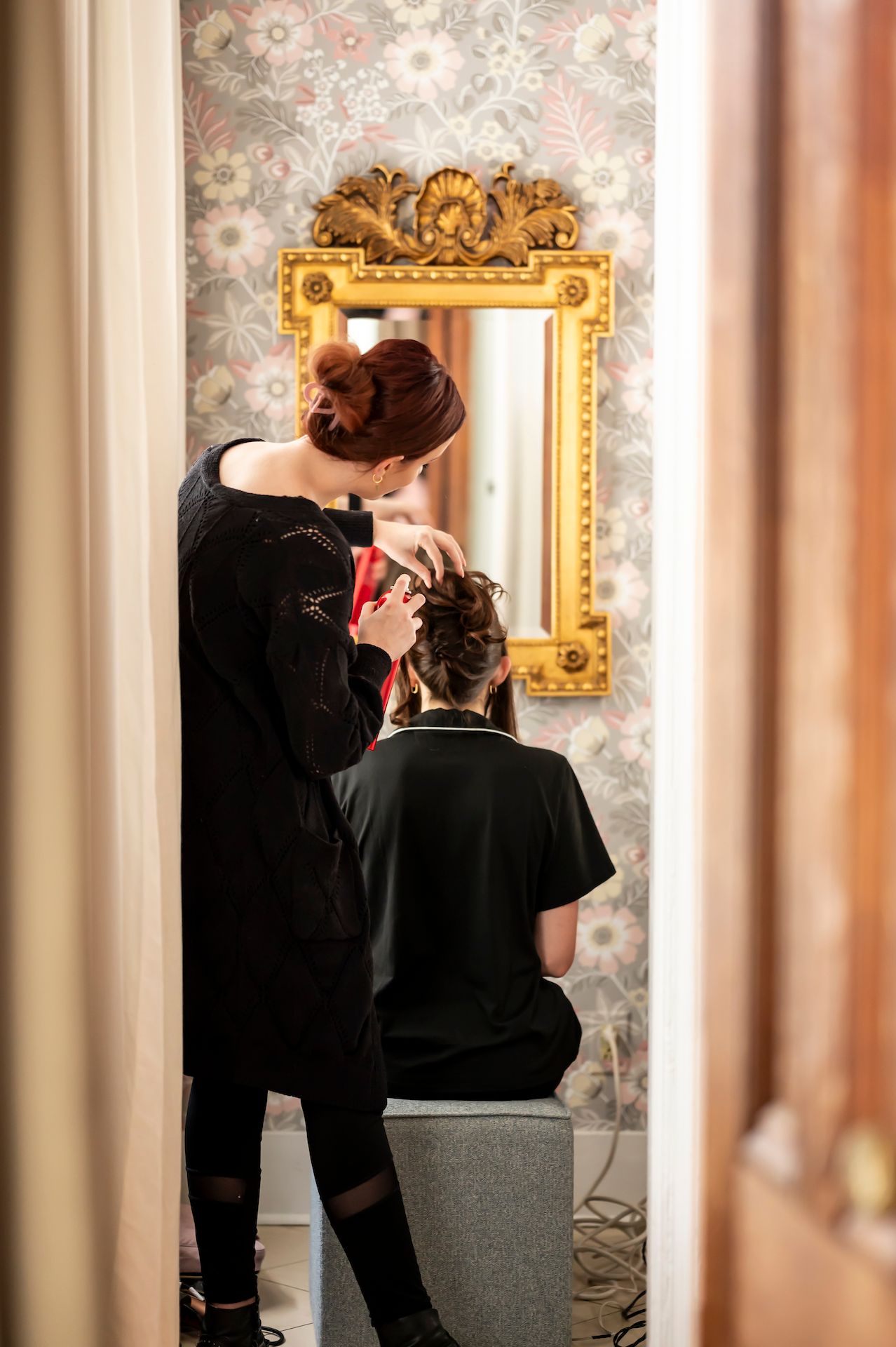 Hairstylist working on client's hair in front of an ornate gold mirror with floral wallpaper background.