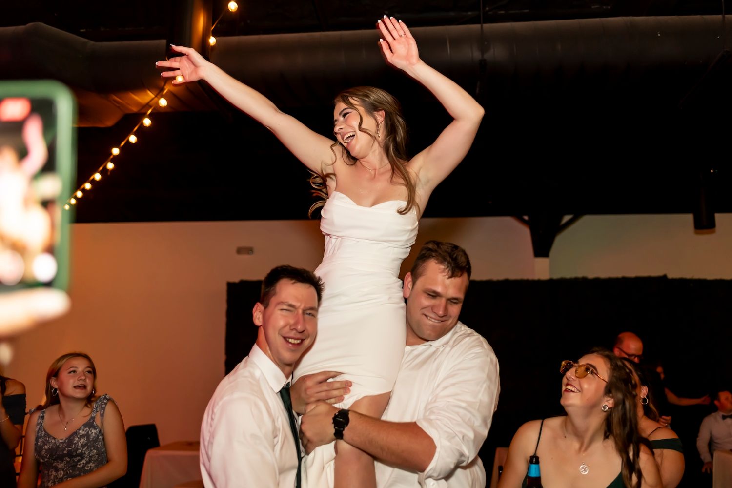 Celebratory dancing and lifting at a wedding reception with string lights overhead and joyful guests.