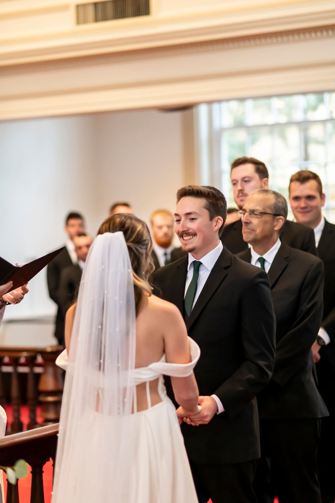 A couple exchanges vows during their wedding ceremony in a church with white columns and red pews.