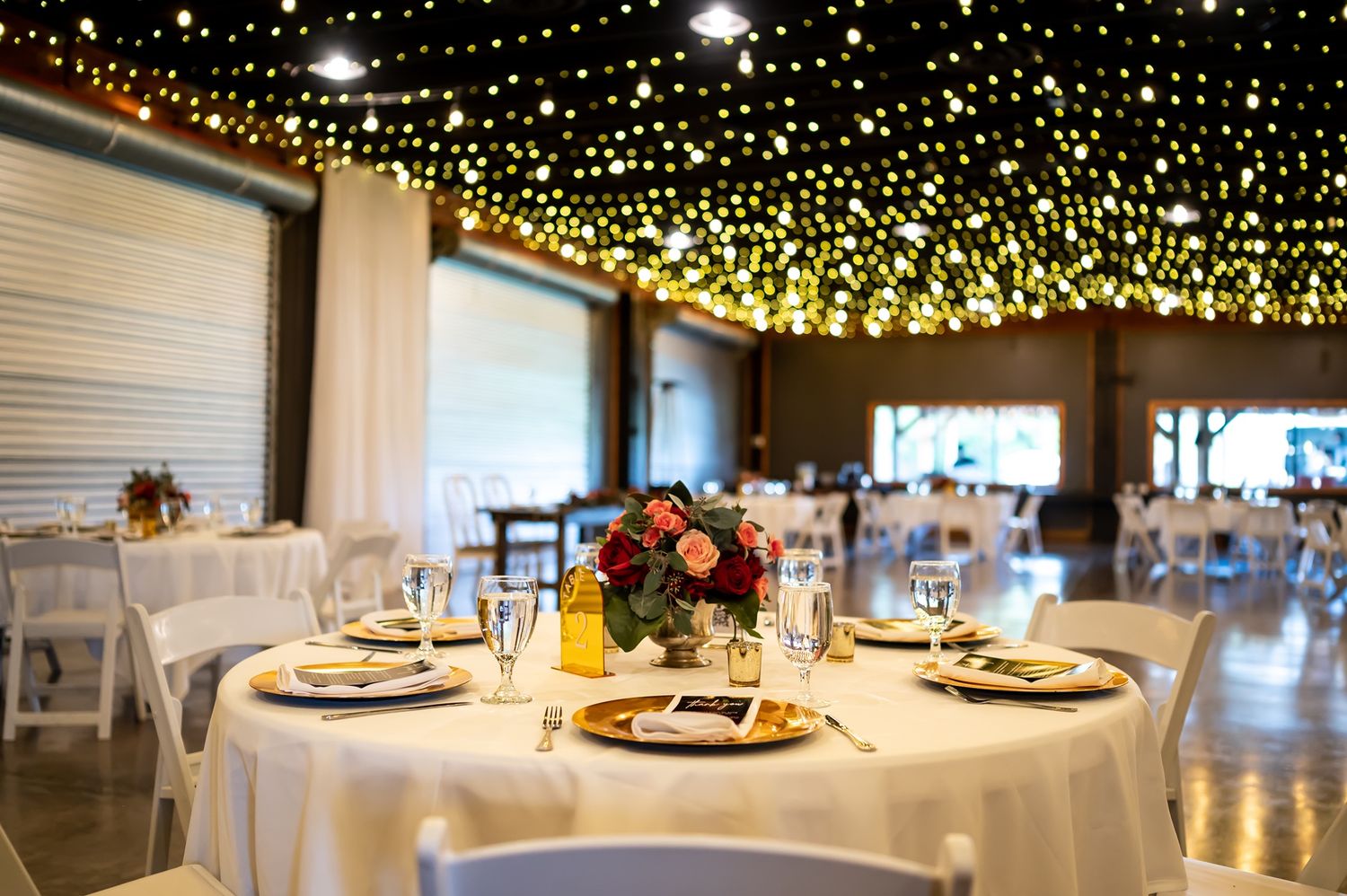 Round dining table set with place settings under warm string lights and floral centerpiece in an event space.
