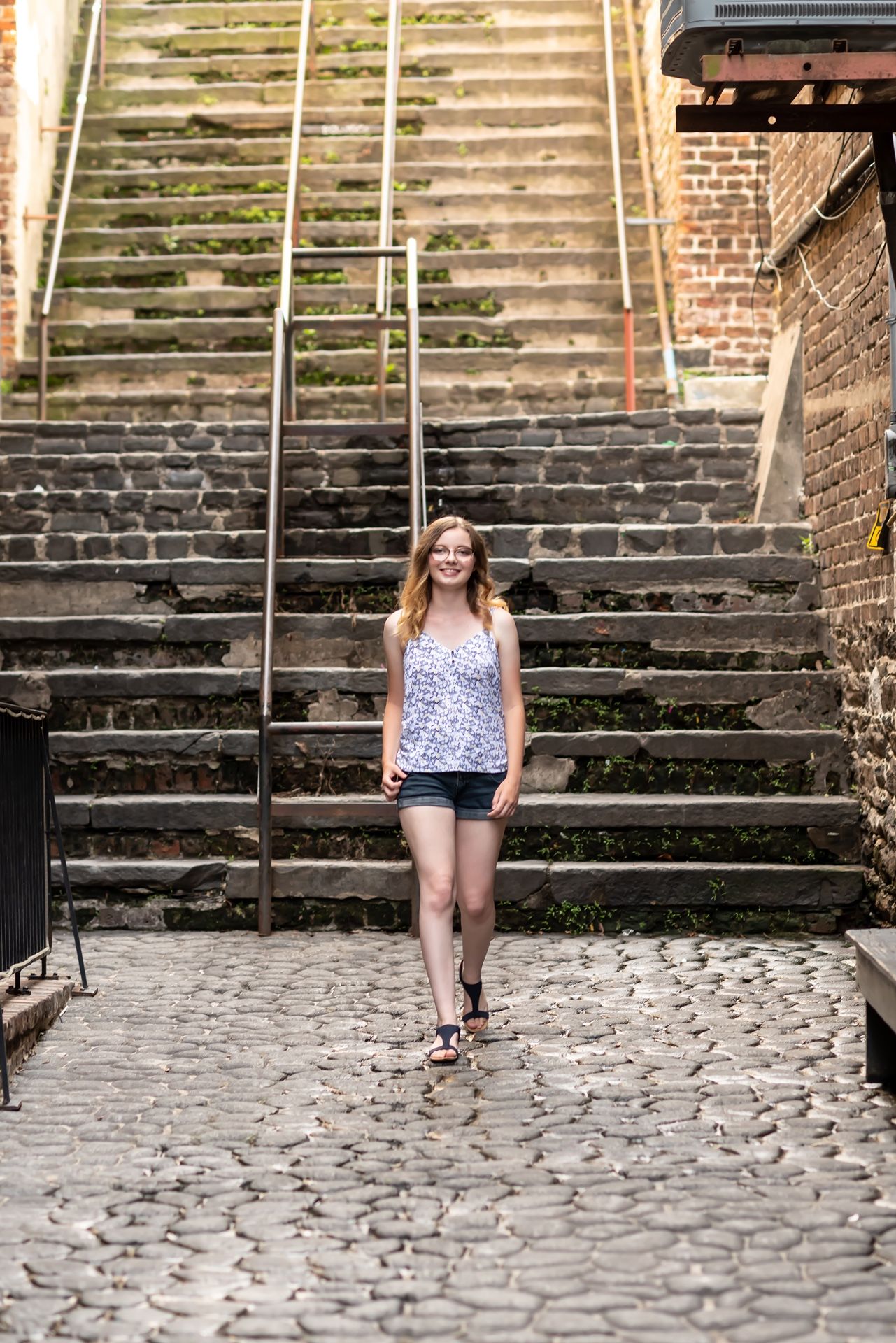 Someone stands at the bottom of historic stone steps with metal handrails against a brick building on a cobblestone path.