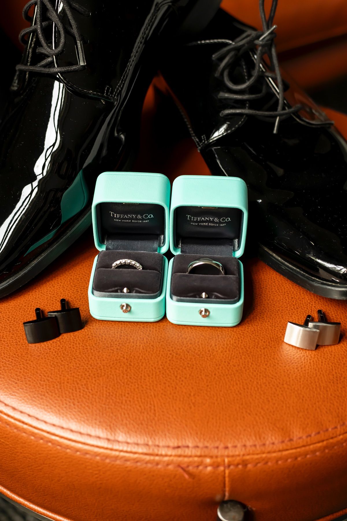 Two Tiffany & Co turquoise ring boxes with wedding rings beside black dress shoes on brown leather surface.