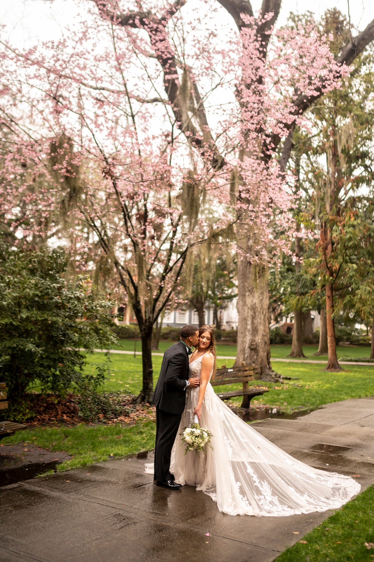 A romantic springtime wedding couple stands beneath blooming cherry blossoms in a lush garden setting.