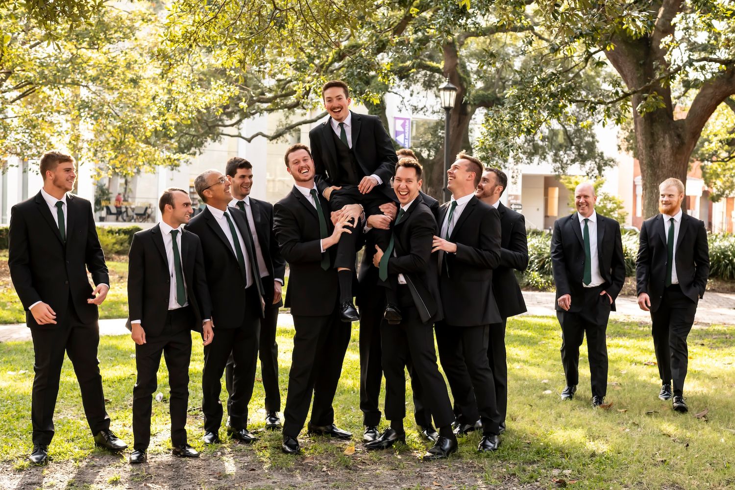 Groomsmen in black suits pose playfully for a group photo on a sunny lawn with trees in the background.
