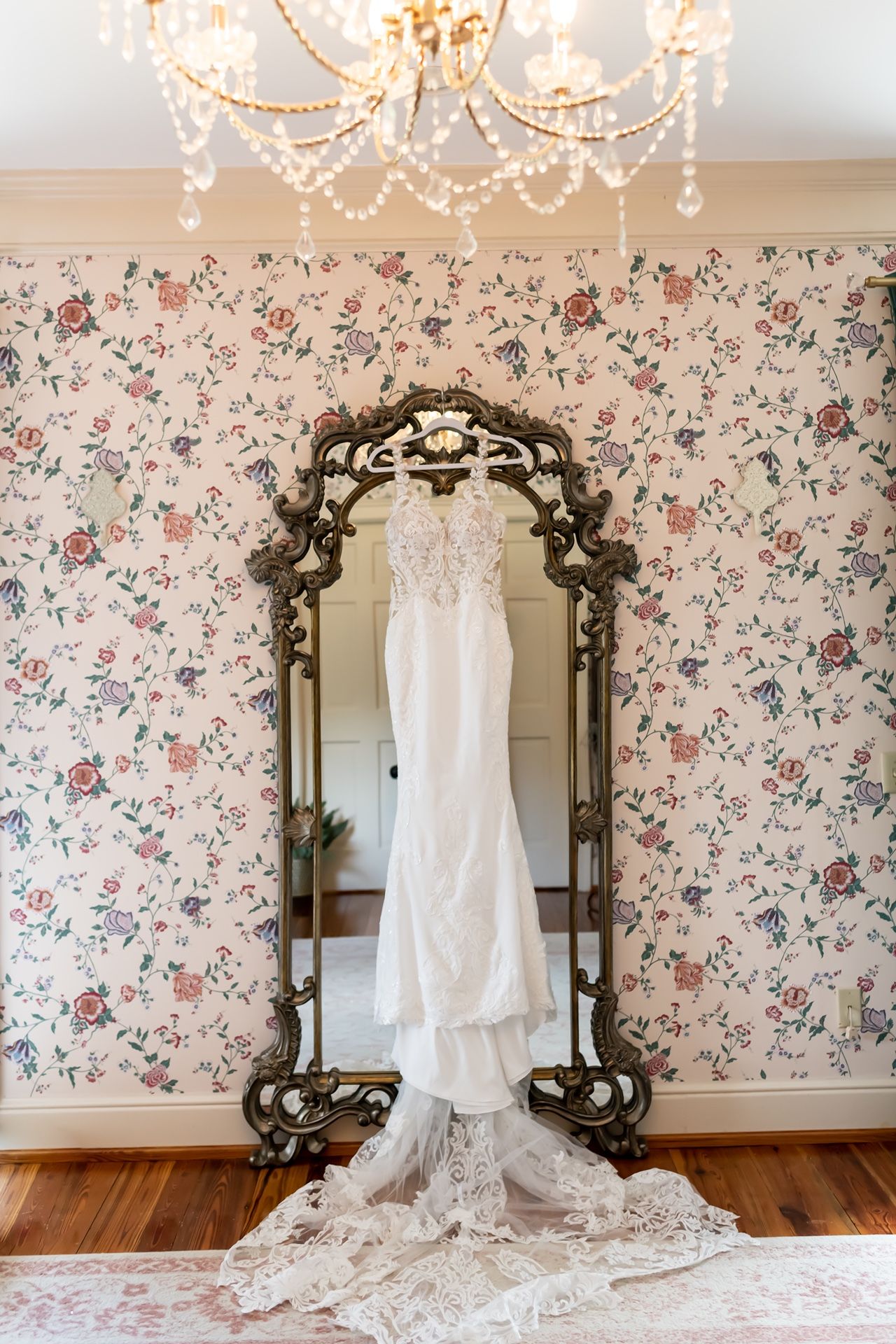 Elegant lace wedding dress hangs from ornate vintage mirror against floral wallpaper with crystal chandelier above.