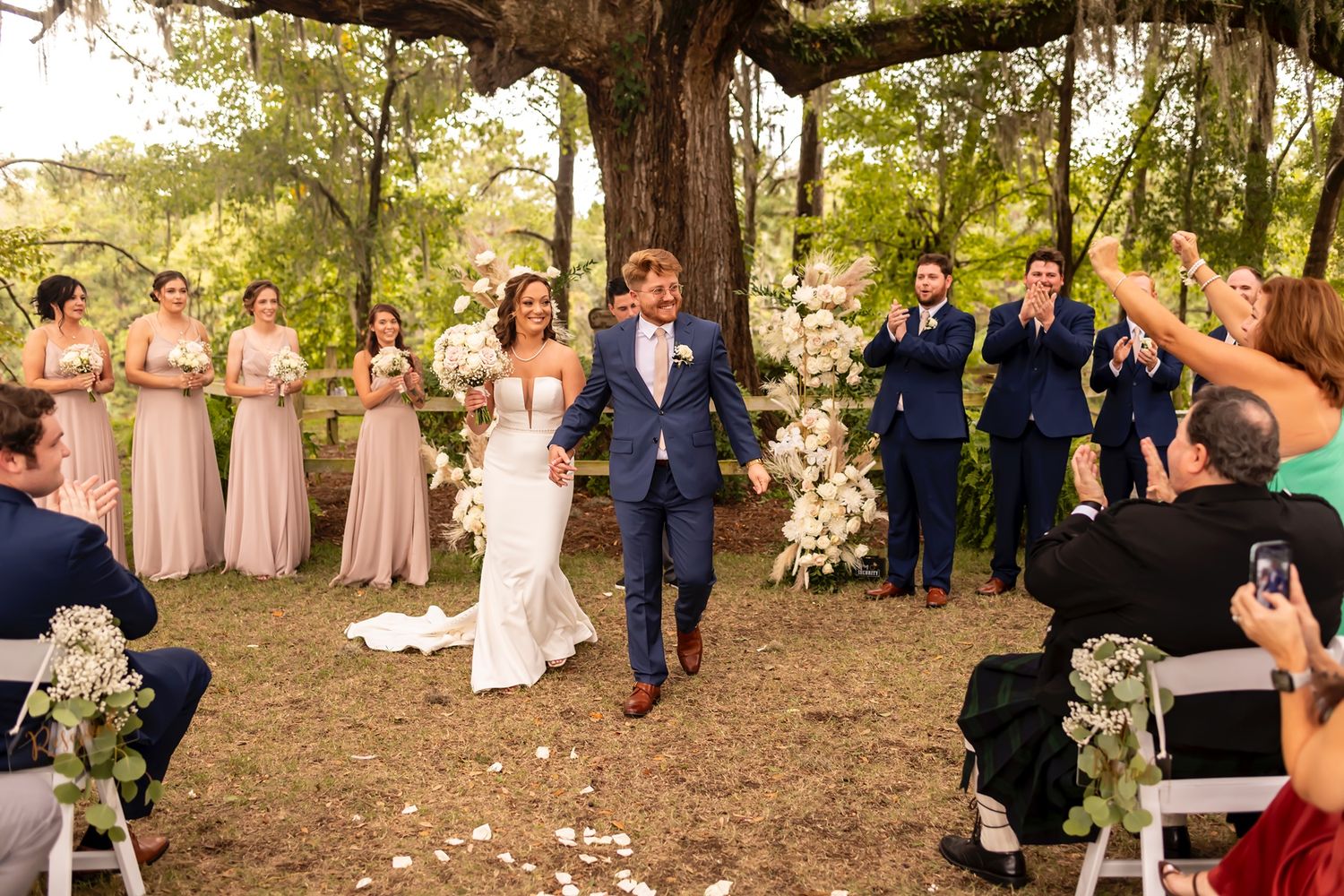 Wedding party lined up for group photos outdoors at sunset in a wooded garden setting.