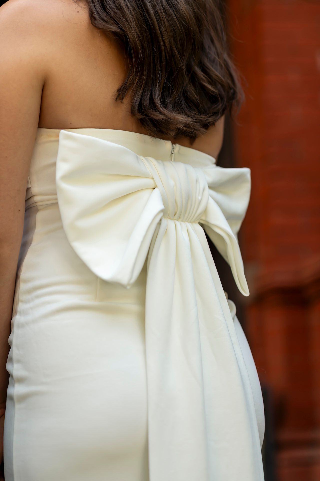 A white formal dress featuring an elegant oversized bow detail in the back against a red-brown background.
