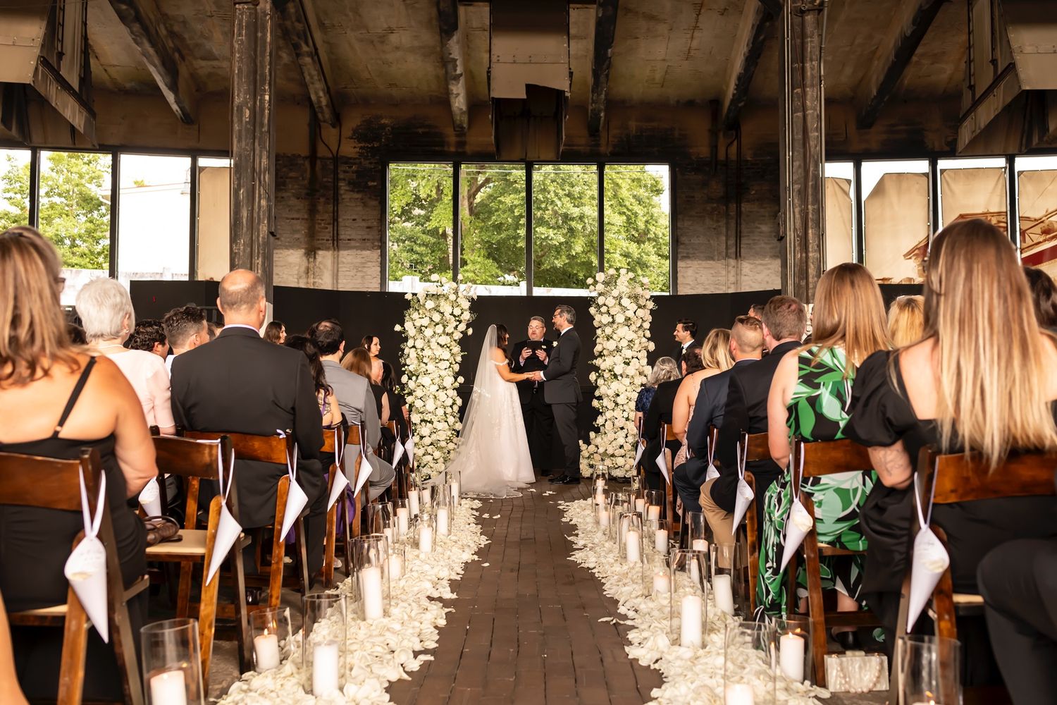 A beautiful indoor wedding ceremony with a white floral arch and rose petals lining the aisle as guests watch from wooden chairs.