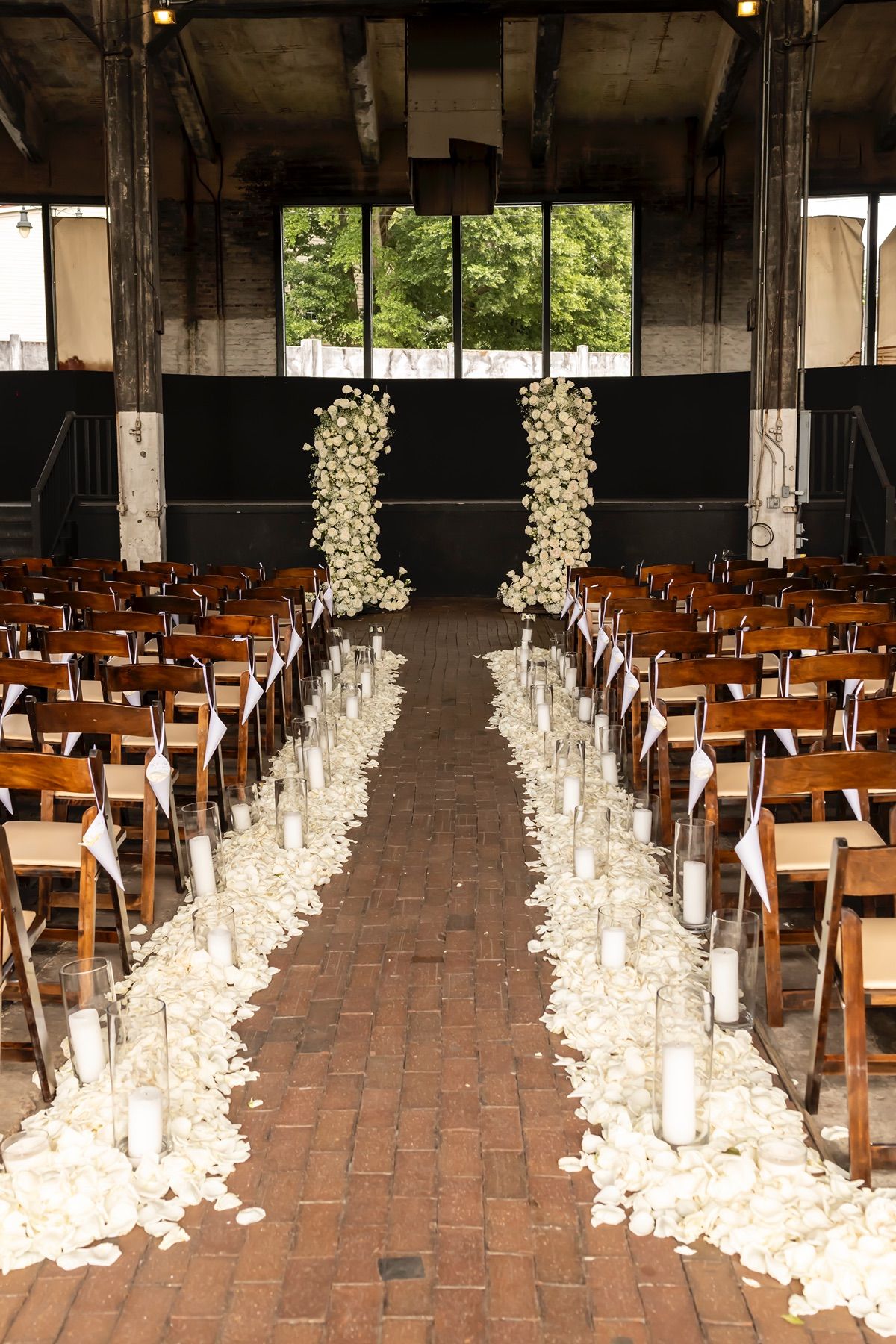 Wedding ceremony aisle decorated with white flower petals and candles lined by wooden chairs in an industrial venue.