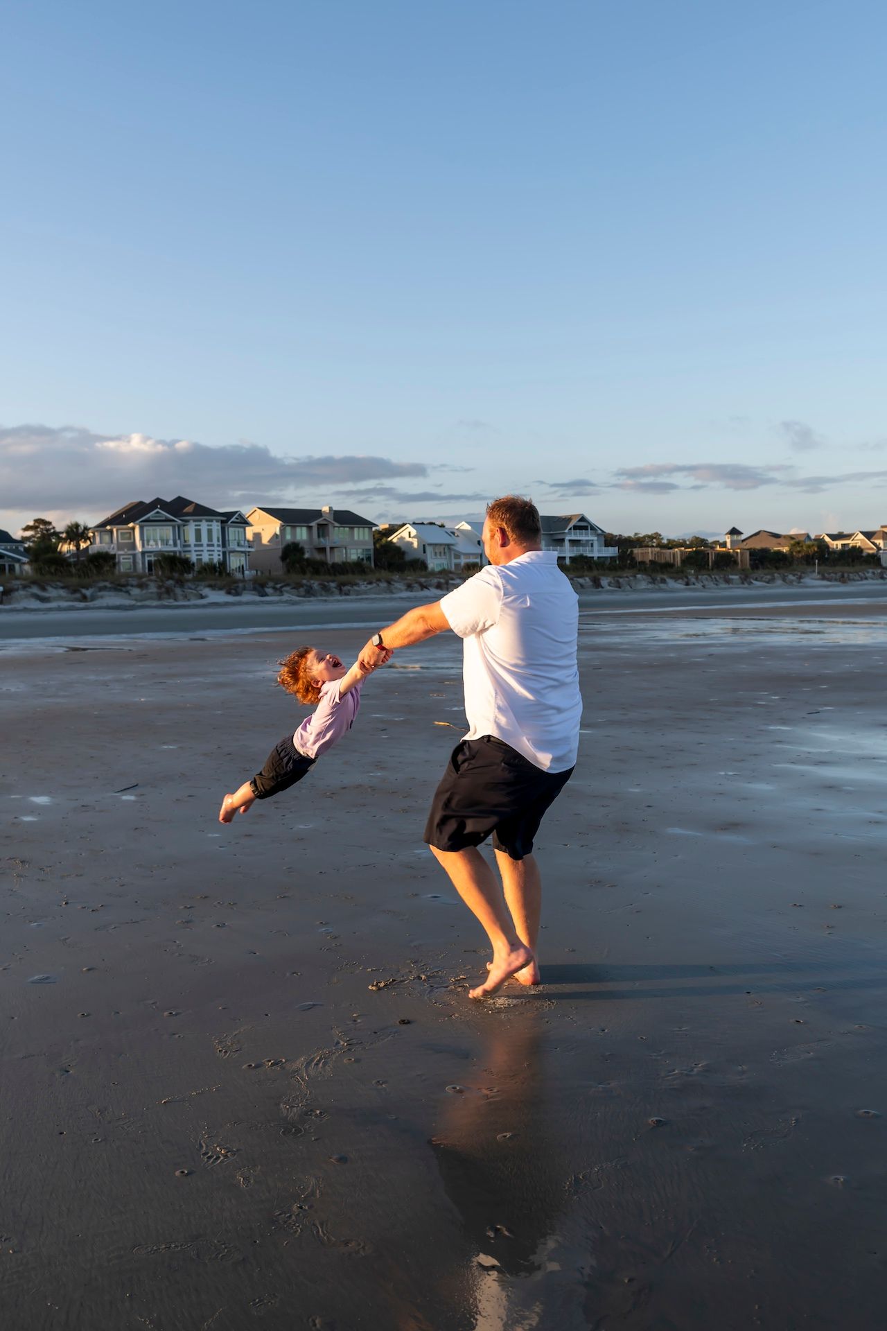 Figures play and jump on a wide empty beach at dusk with beach houses visible in the distance.