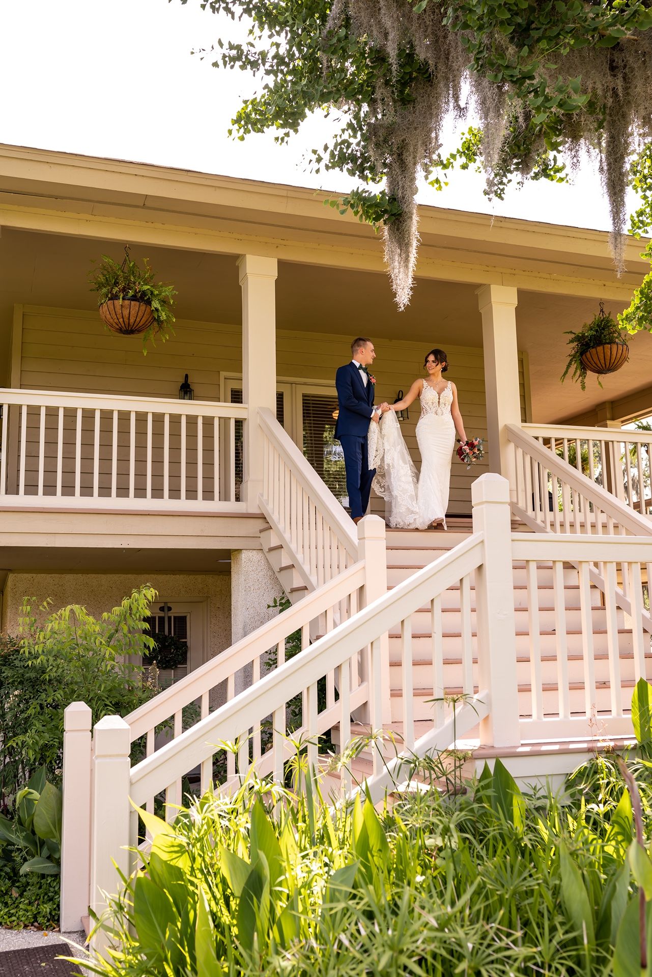 White Southern-style plantation house porch with steps surrounded by lush tropical foliage and hanging ferns.