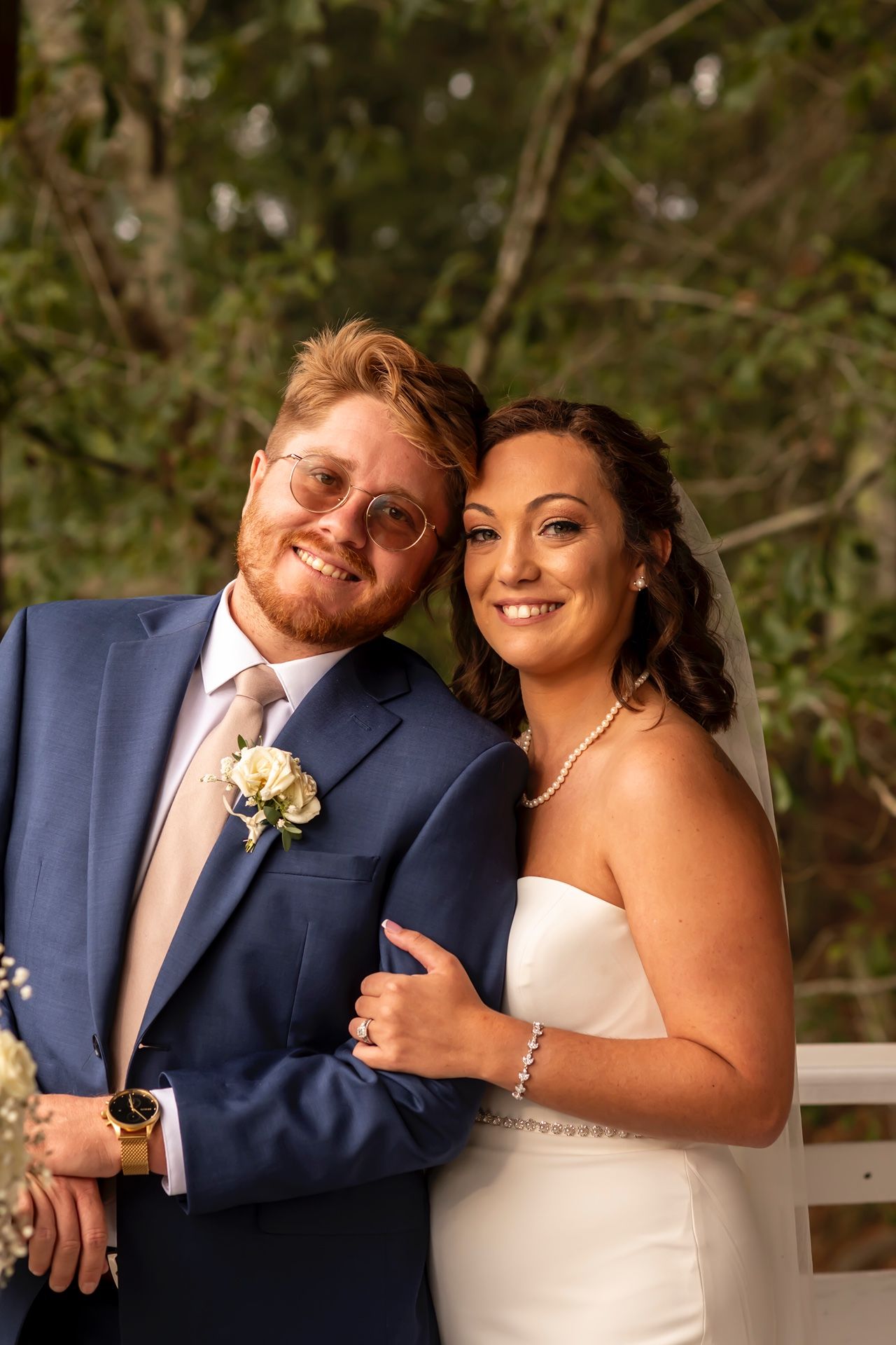 A wedding couple in formal attire poses outdoors, bride in white dress and groom in navy suit with blush tie and boutonniere.