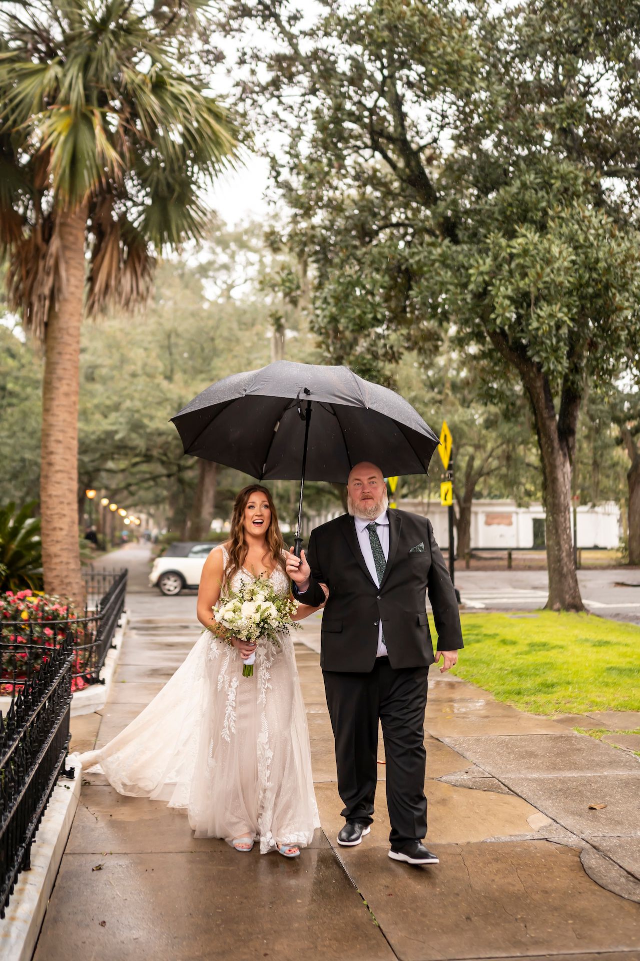 A couple walks along a rainy sidewalk under an umbrella with palm trees and greenery in Charleston, South Carolina.