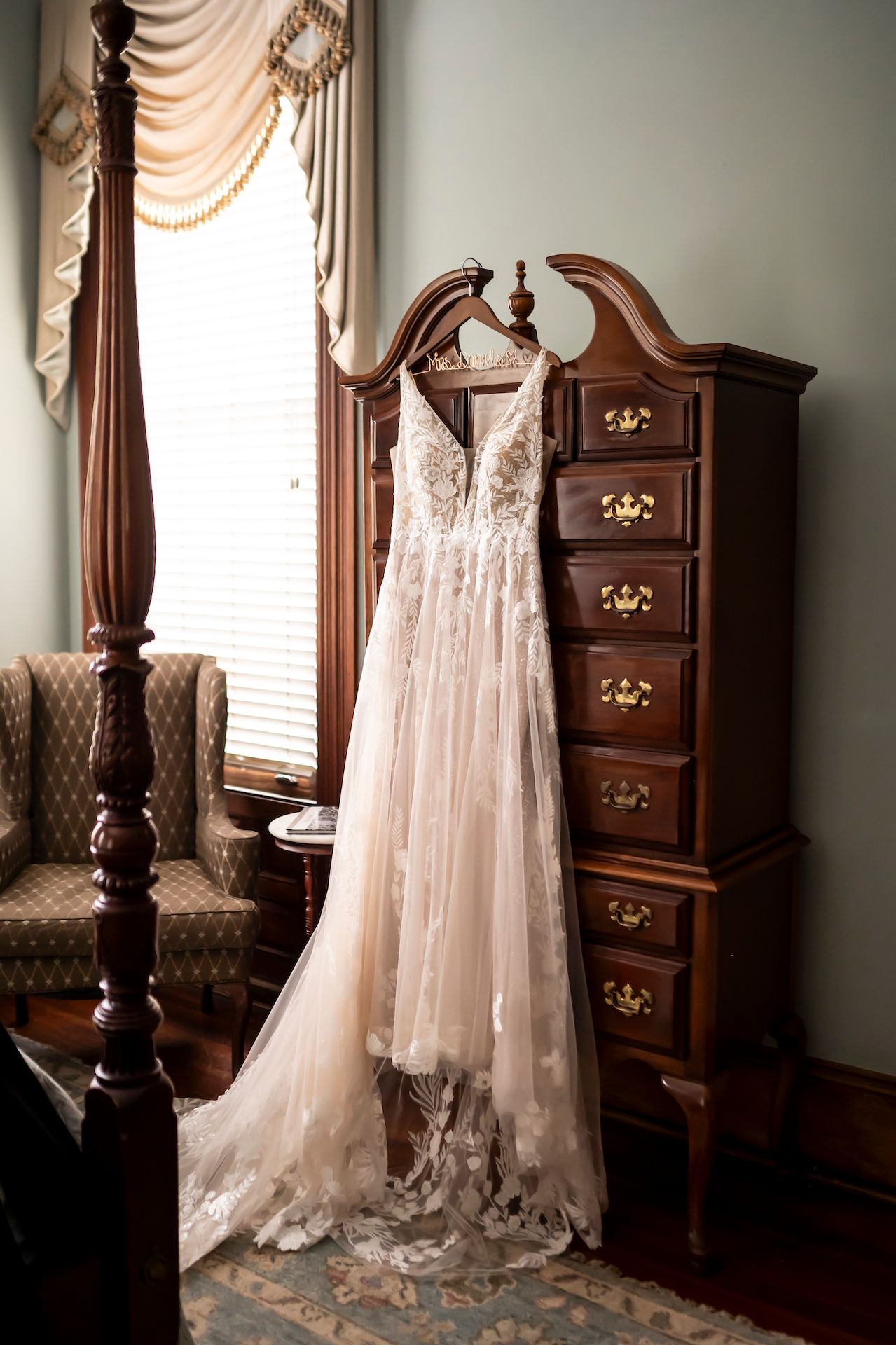 A delicate lace wedding gown hangs from an ornate wooden bed post next to an antique tall dresser by a draped window.