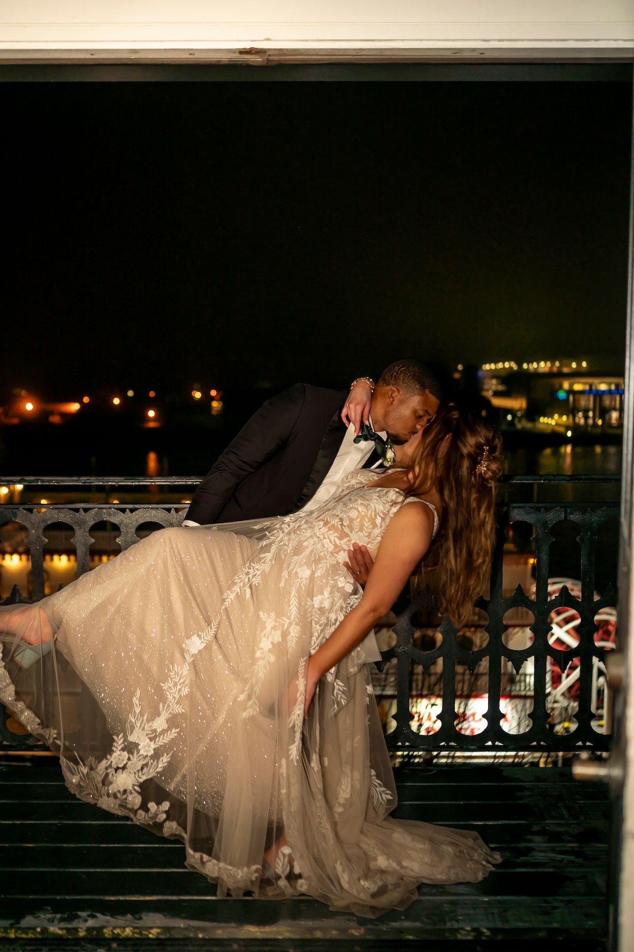 Couple shares romantic kiss in dip pose on balcony at night with lace wedding dress and city lights in background.