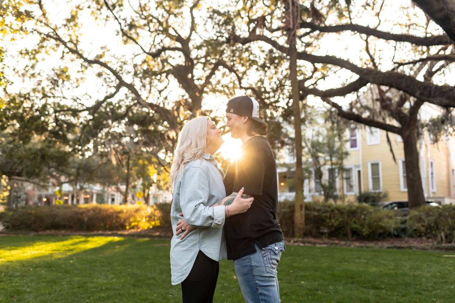 Two figures embrace in a park at golden hour with sunlight streaming through tree branches and a building in background.