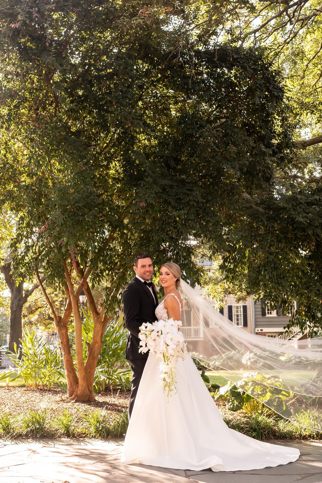 Newlyweds pose together under lush trees in garden setting as bride's long veil flows in breeze.