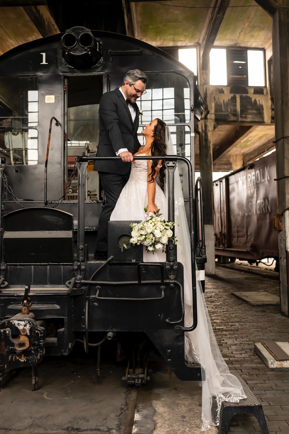 Couple takes romantic wedding photos on vintage black steam train engine number 1 with flowing white veil and bouquet.
