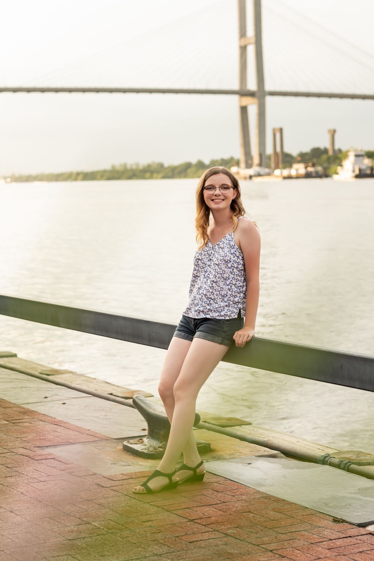 A casual summer outfit and smile while standing at a waterfront dock with a bridge visible in the background.