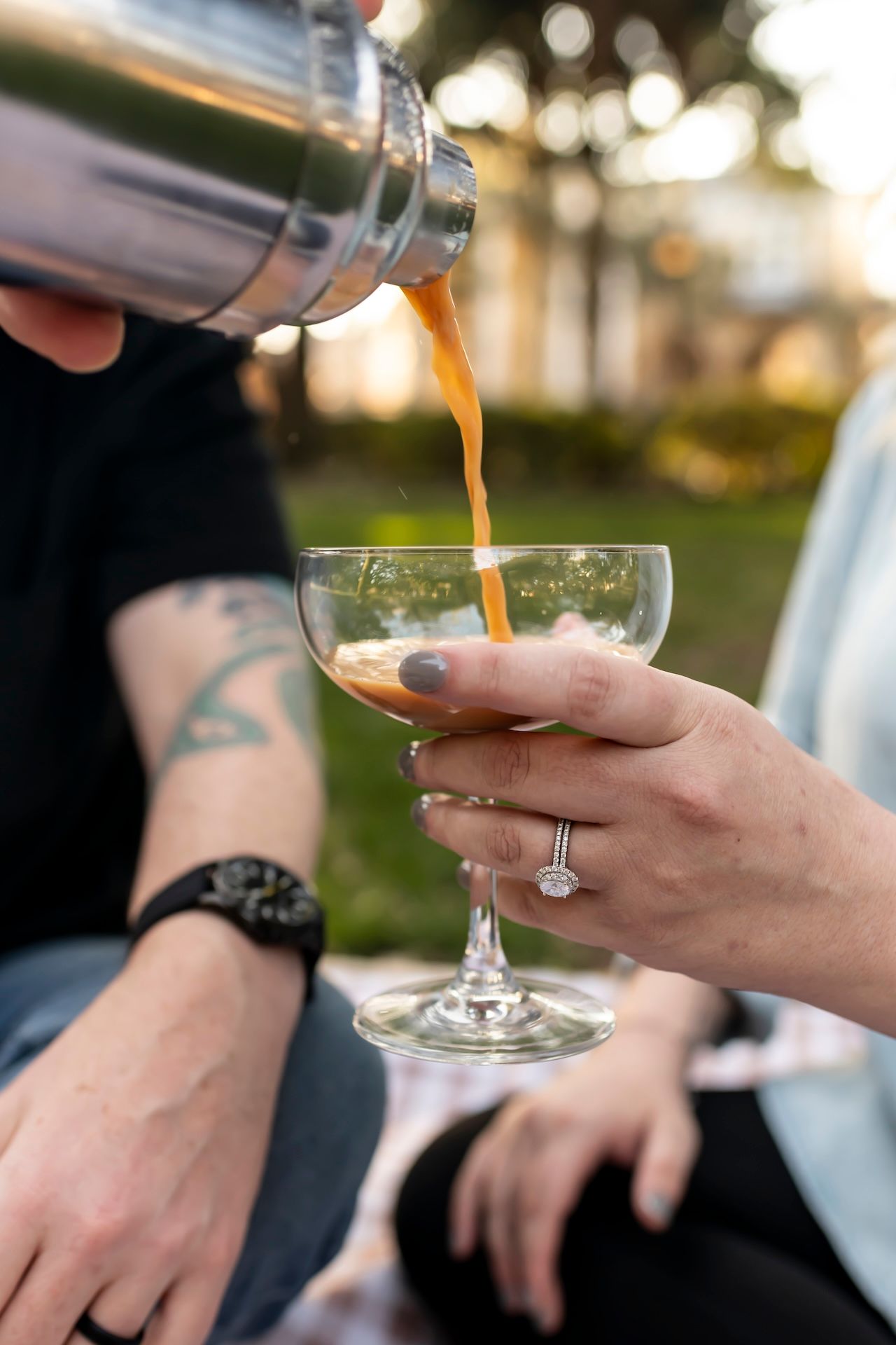 Someone wearing a black sleeve pours a golden cocktail from a shaker into an elegant coupe glass held by another hand.