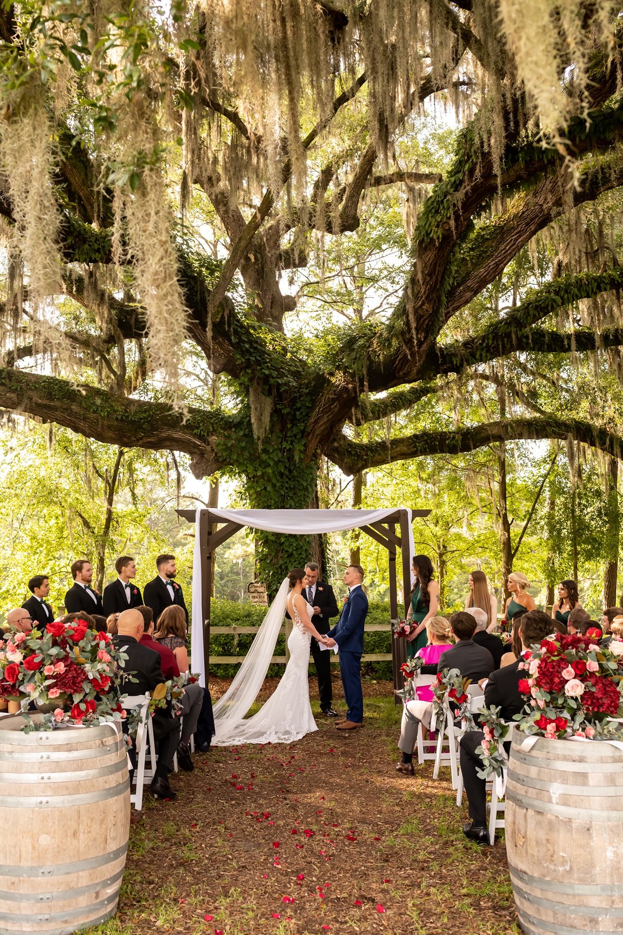 Outdoor wedding ceremony under a majestic oak tree draped with Spanish moss and decorated with rustic wine barrels and red roses.