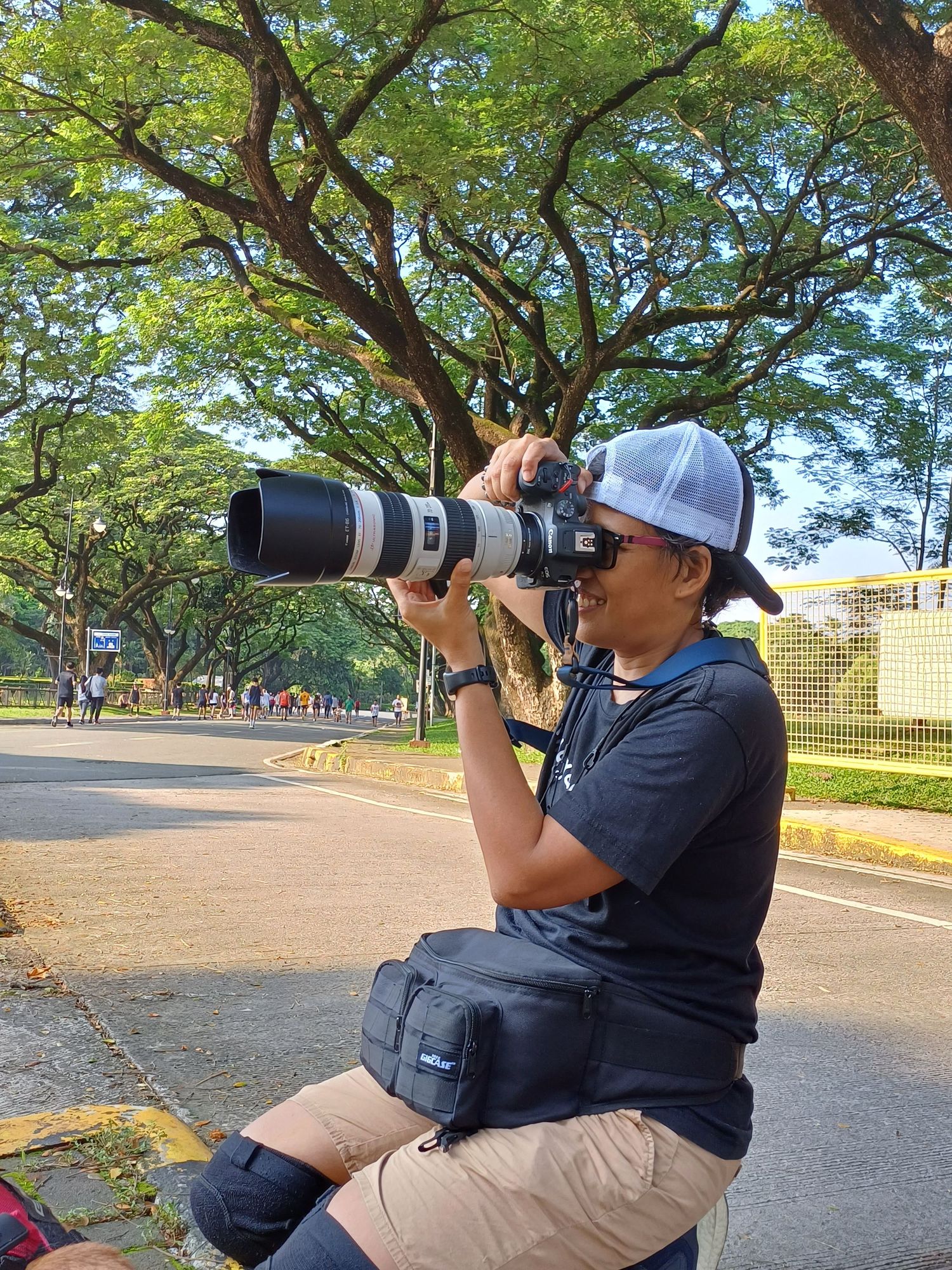 Photographer with professional camera and telephoto lens shooting outdoors under large tree.
