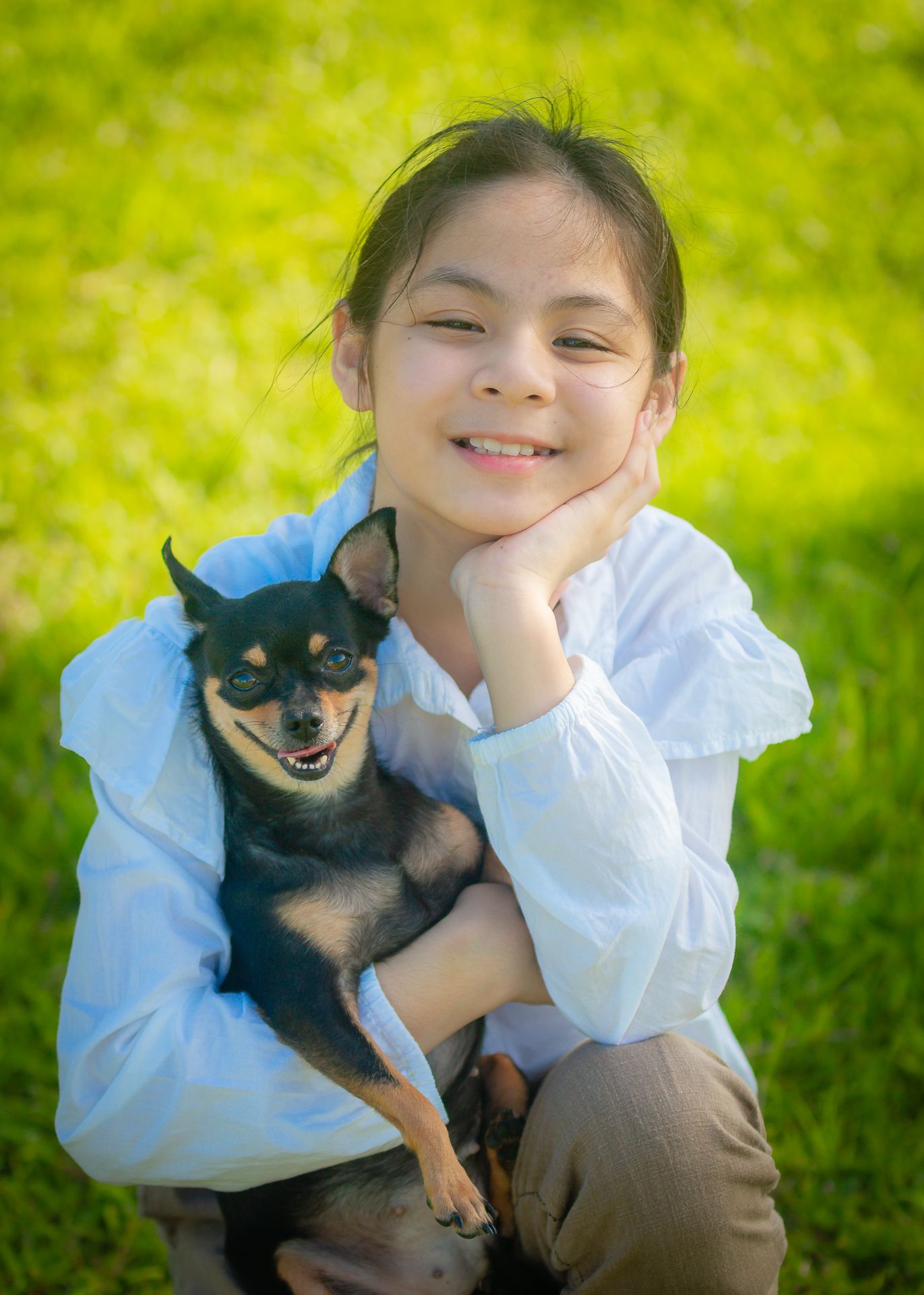 A young person in a white sweater smiles while holding a small black and tan Chihuahua dog on green grass.