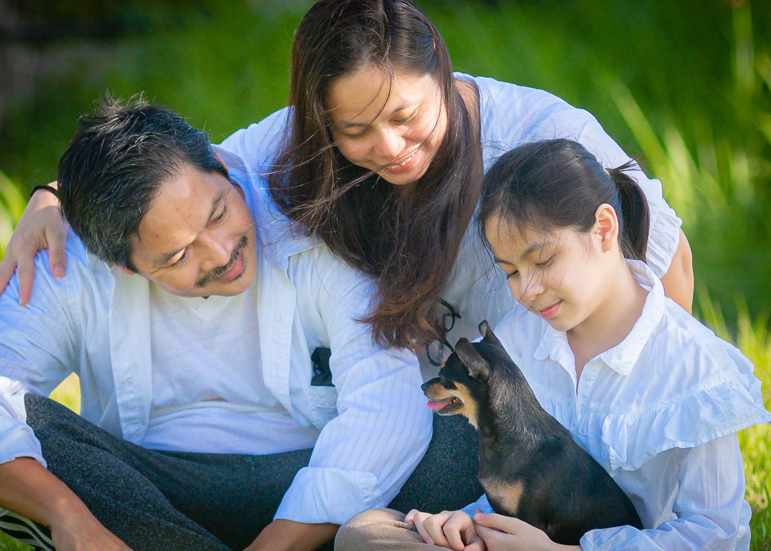 Three people in white shirts smile while petting a small dark-colored dog outdoors in a sunny garden setting.