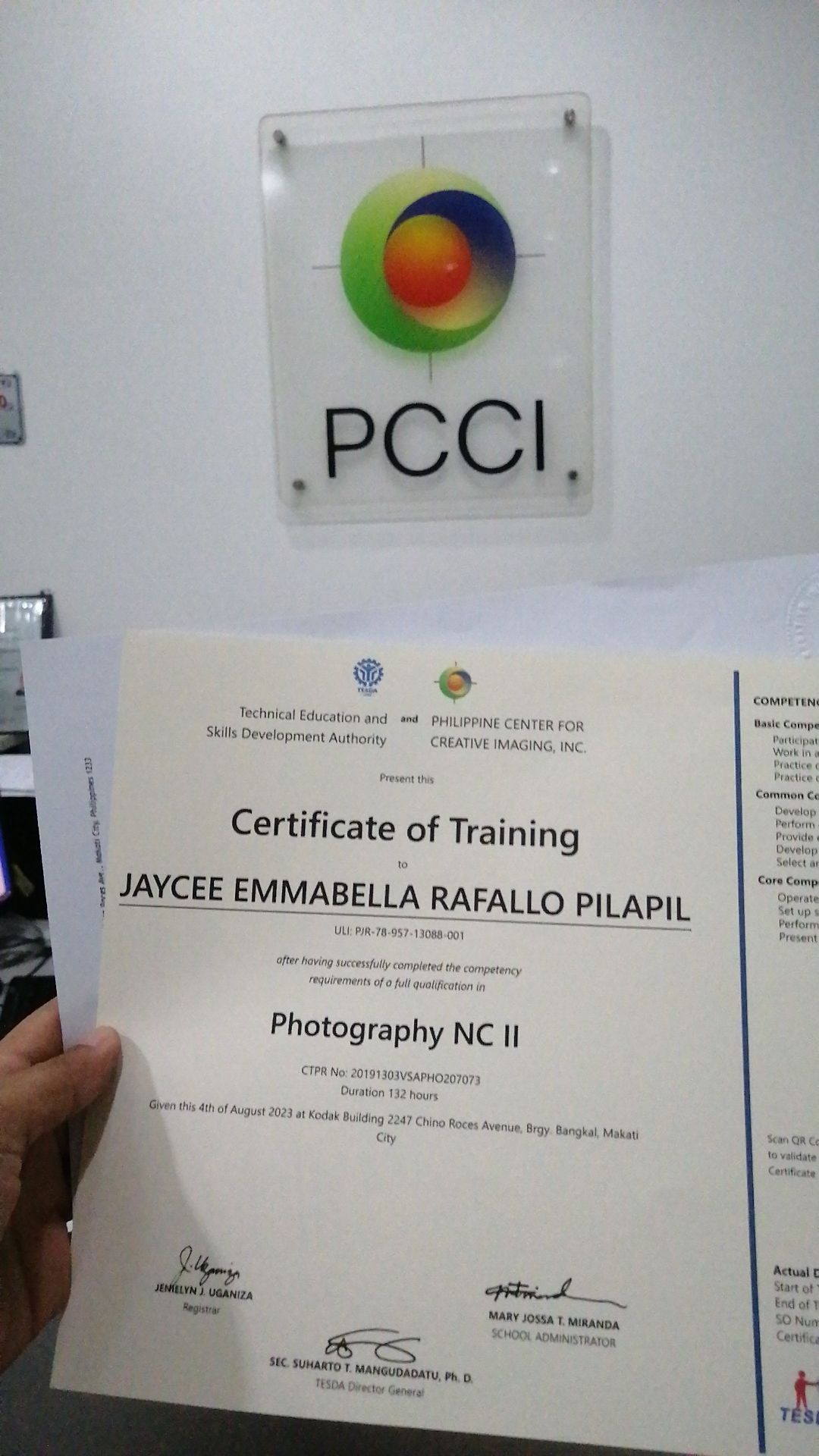 A Photography NC II certificate of training document shown below a PCCI logo mounted on a clear acrylic wall display.