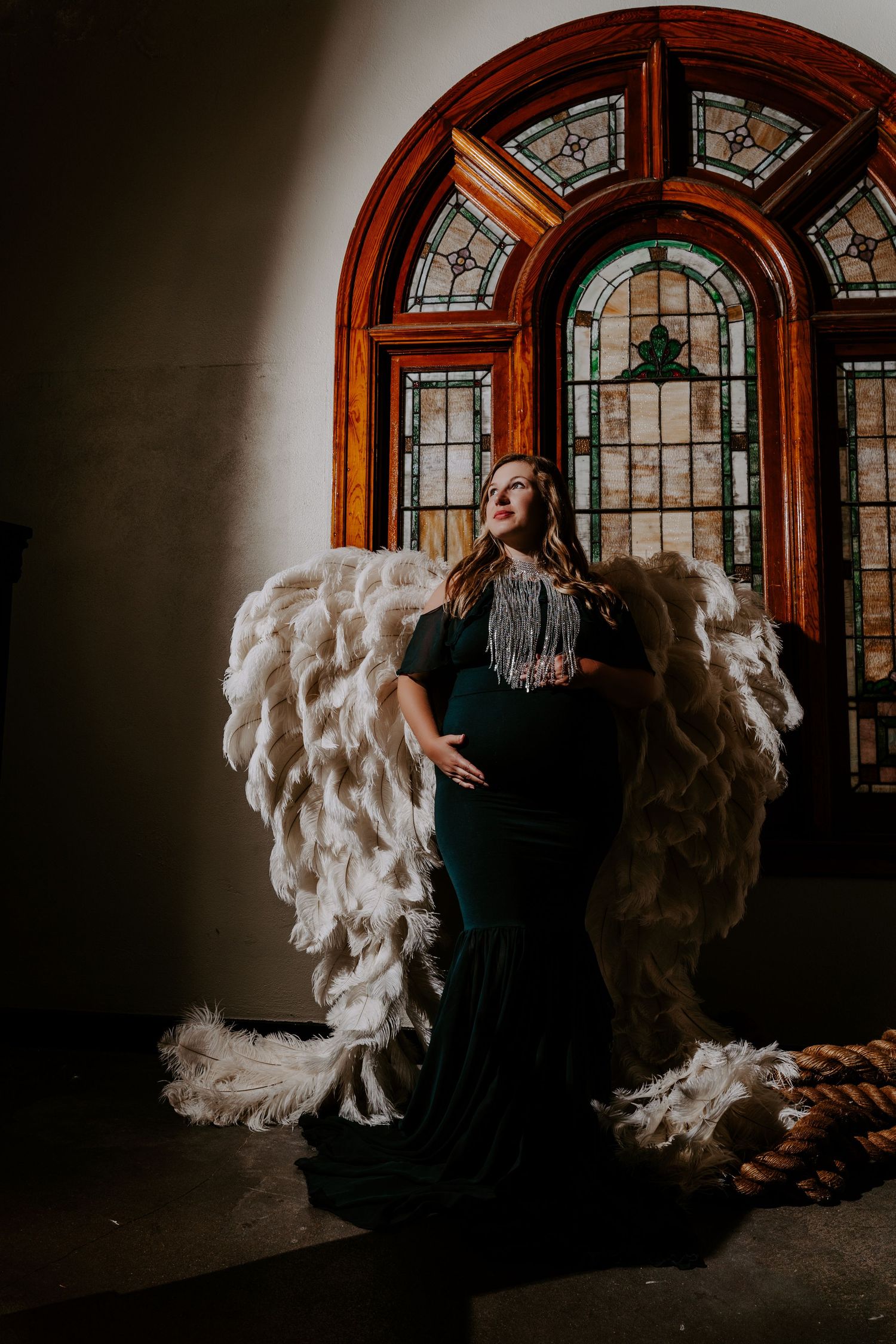 Fashion model wearing black dress and crystal beaded necklace poses in front of arched stained glass window and white wings.