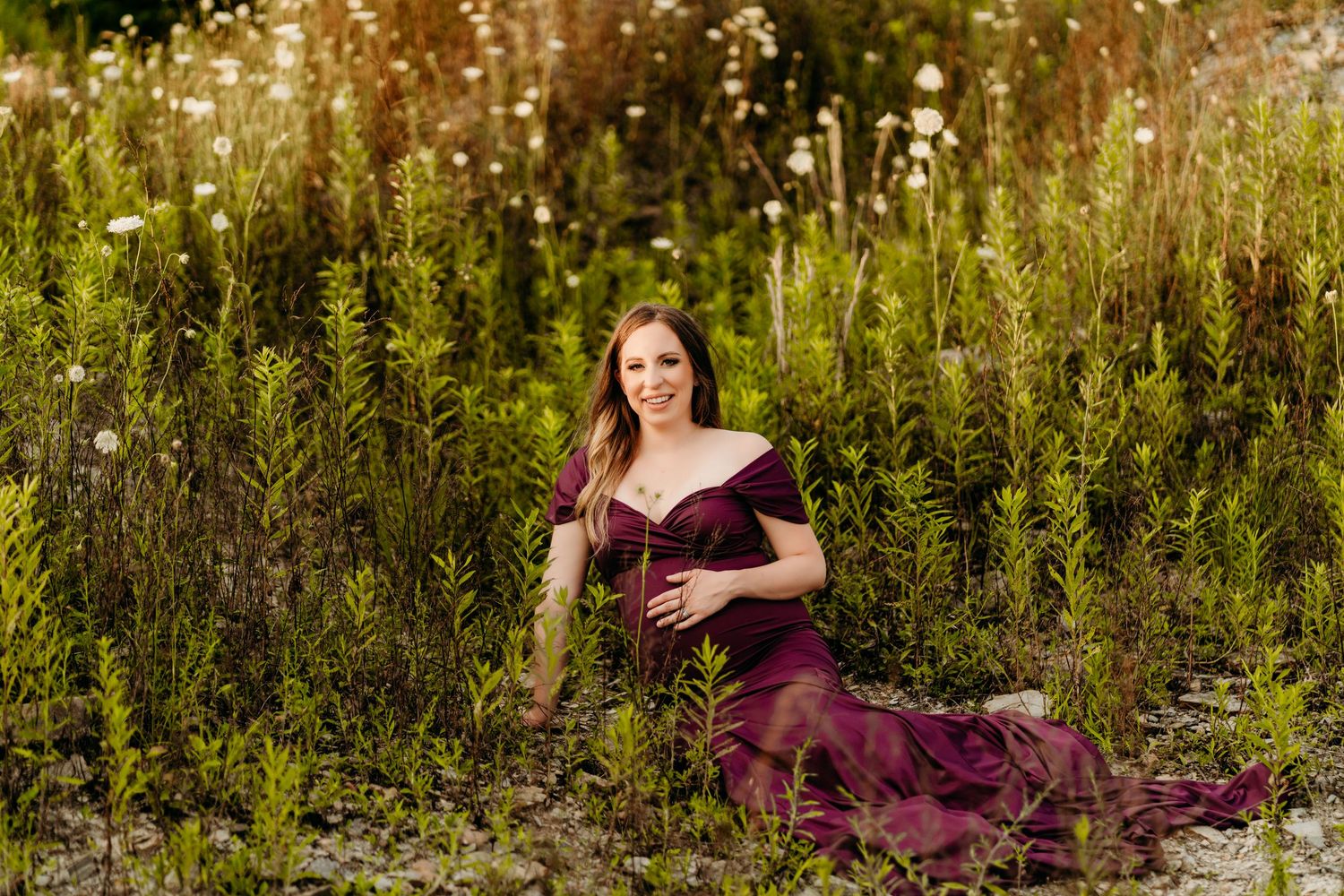 Maternity portrait in a burgundy gown among tall wildflowers at sunset.