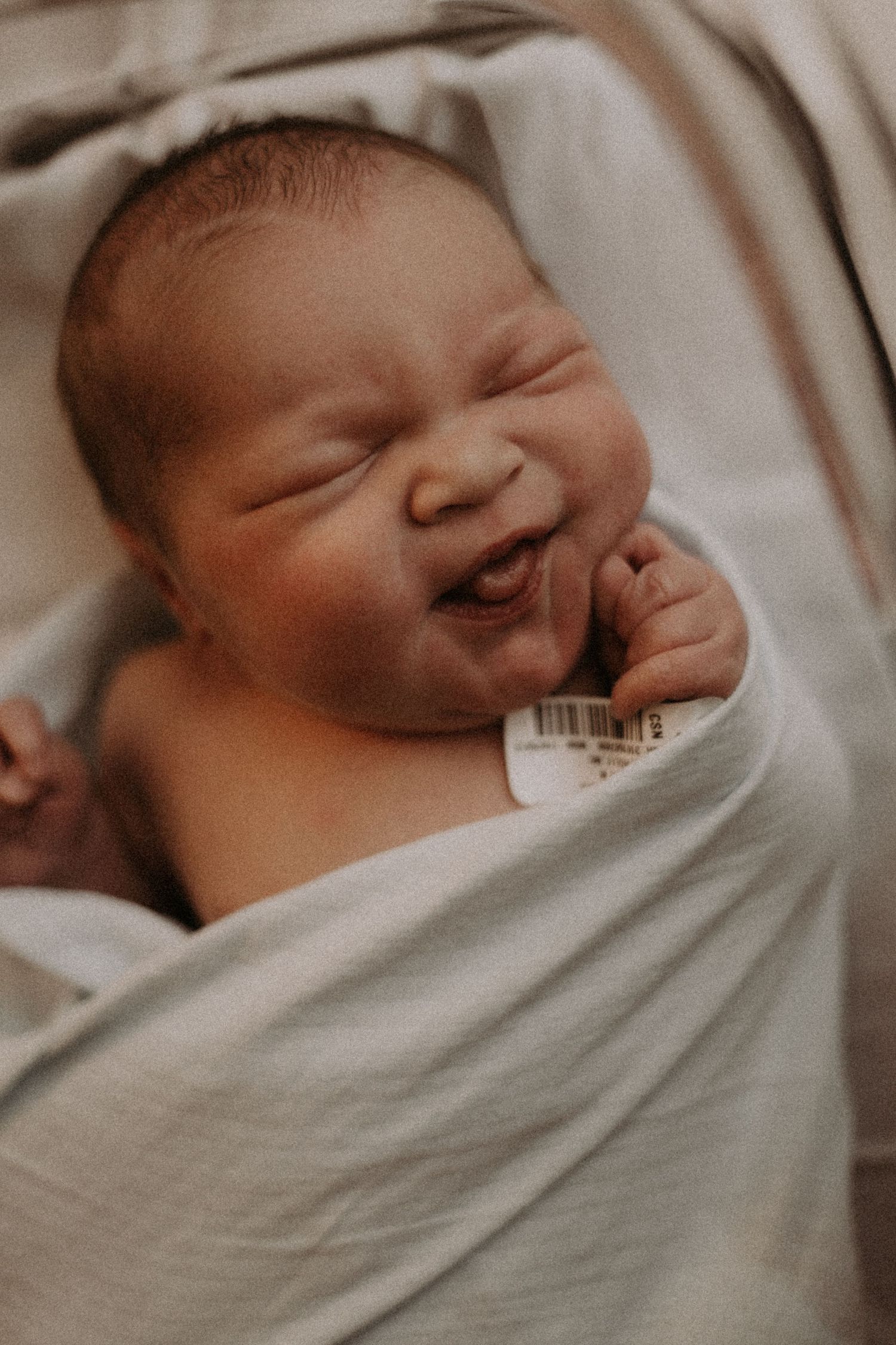 Newborn baby wrapped in soft white blanket shown sleeping peacefully in a sepia-toned photoshoot.