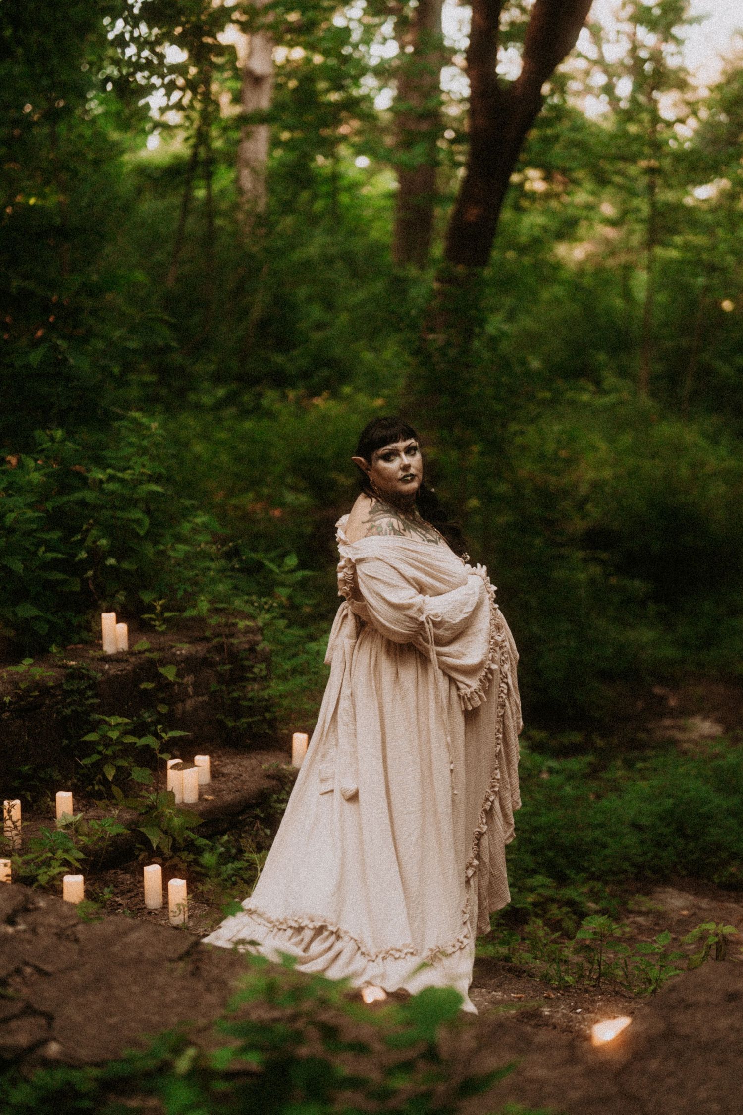 A figure in a flowing cream-colored vintage dress stands in a wooded forest setting illuminated by glowing candles on the ground.