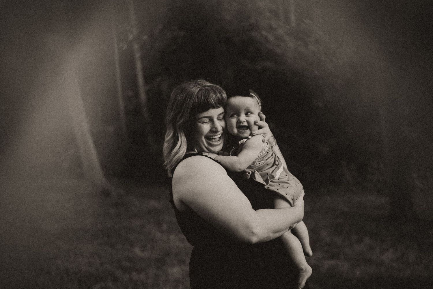 A black and white portrait of a mother and baby smiling together in a dreamy outdoor setting.