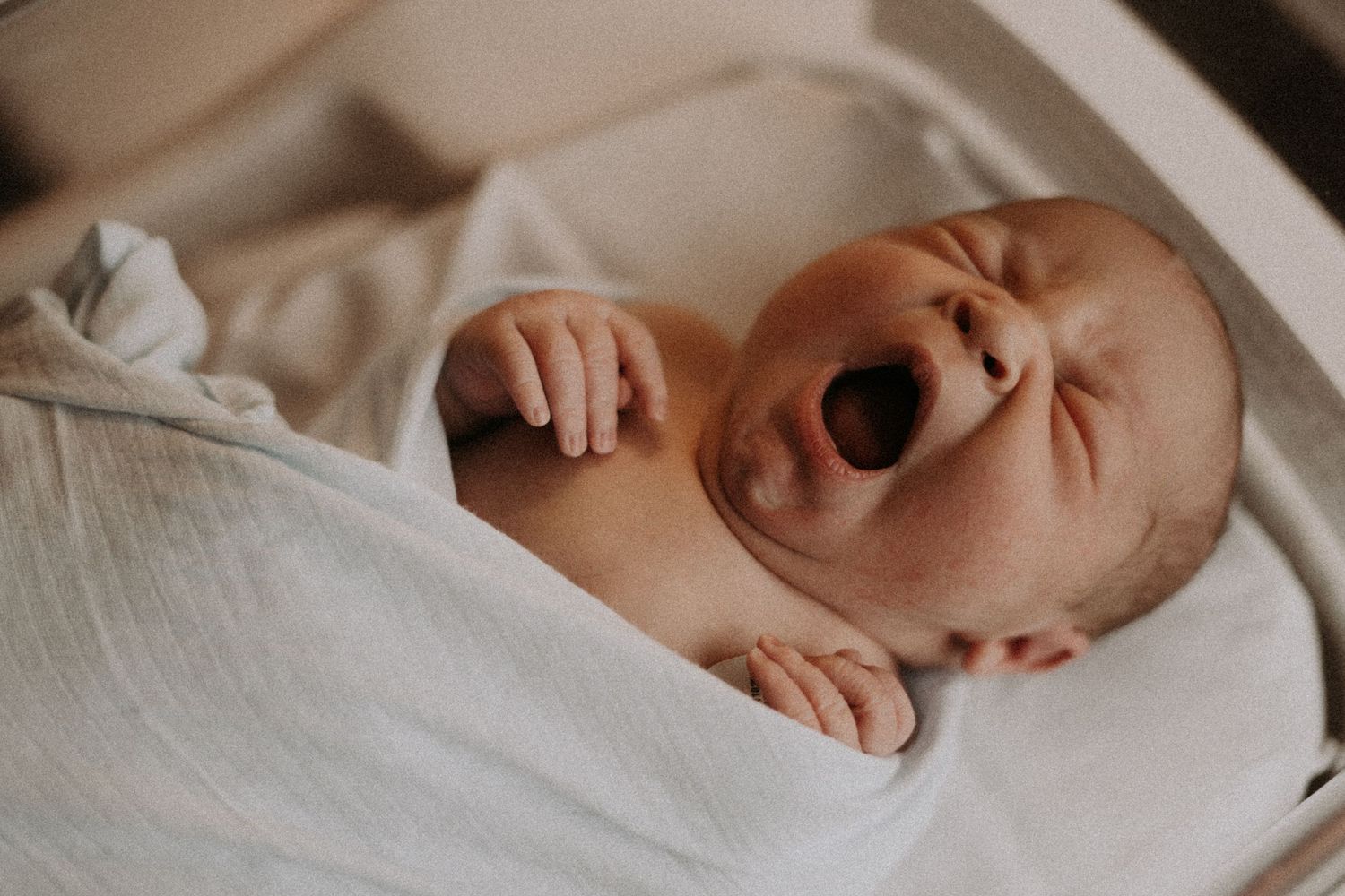 Newborn baby yawning while wrapped in white swaddle blanket.