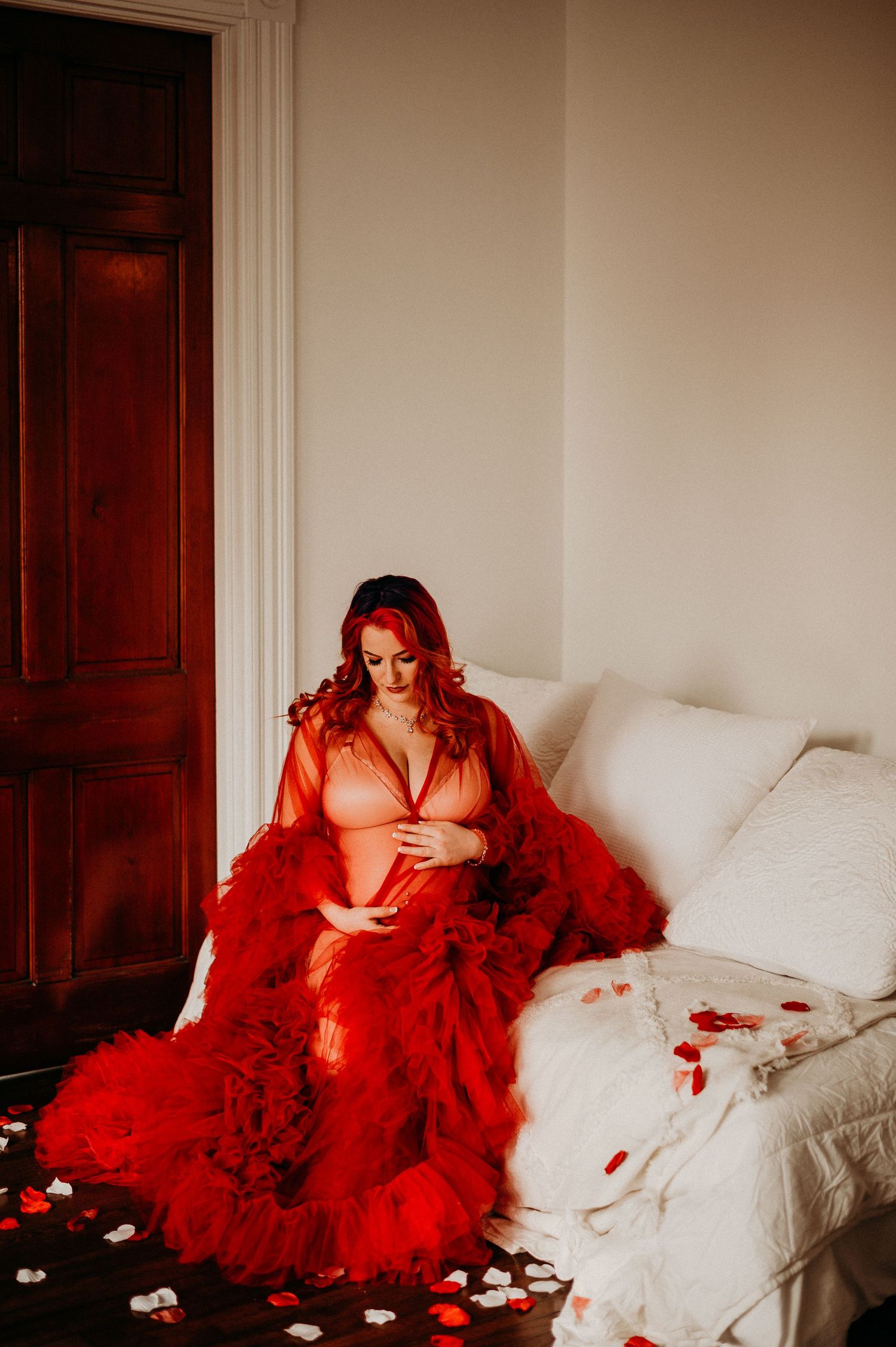 Dramatic red maternity photo with rose petals on white bed.