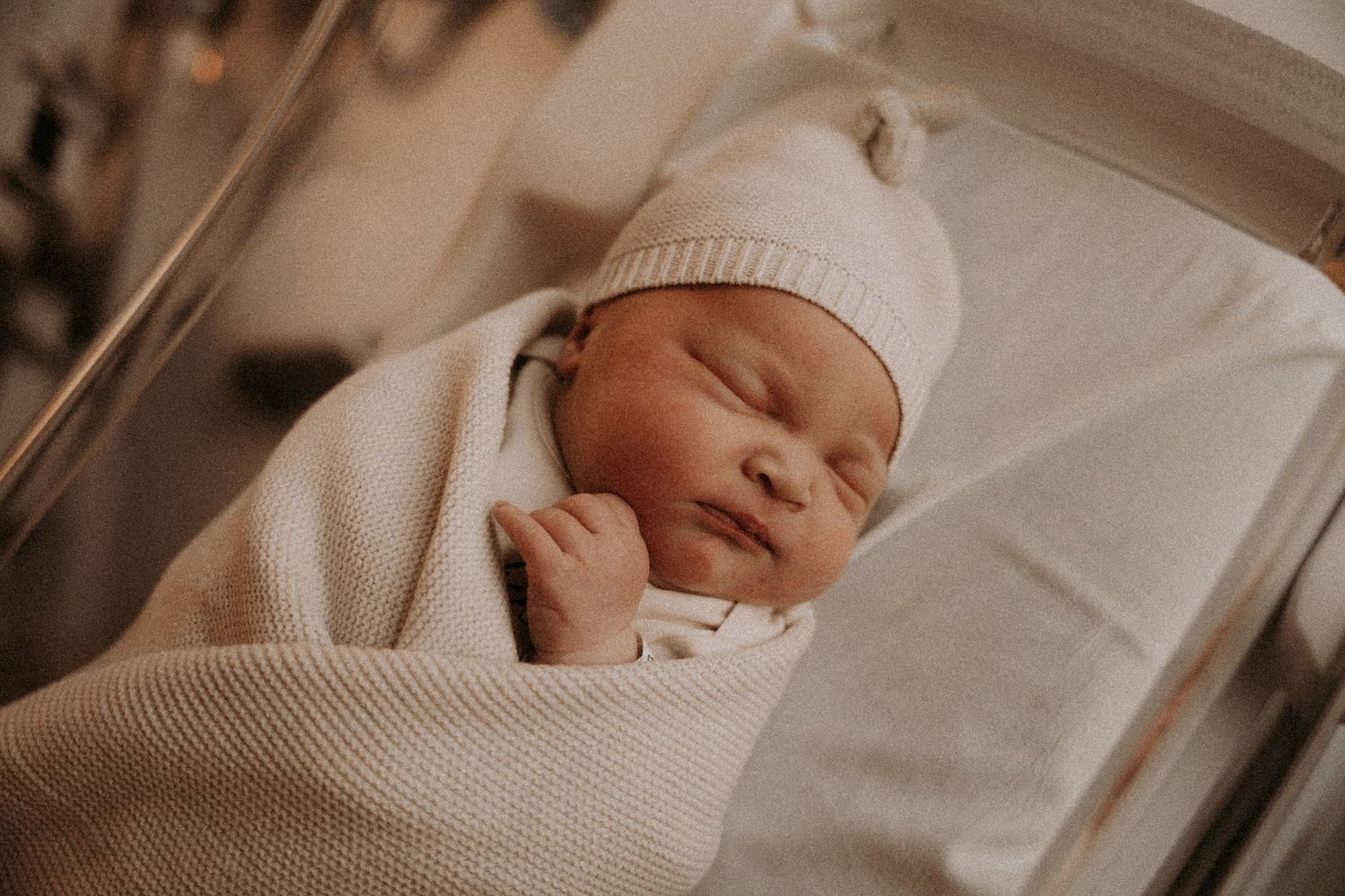 Peaceful newborn swaddled in cream knit blanket and matching hat sleeping soundly.