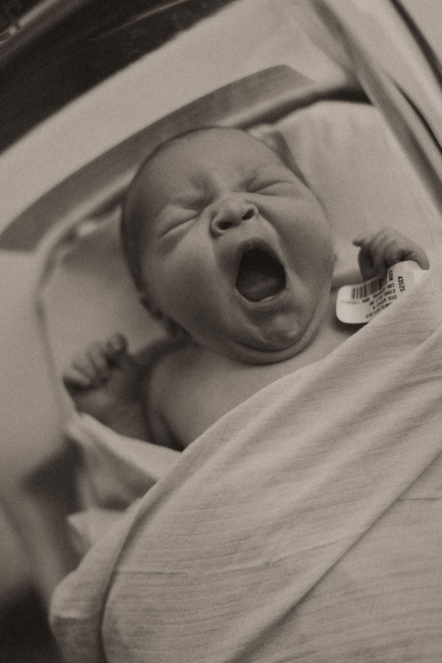 Black and white portrait of yawning newborn baby wrapped in hospital blanket.