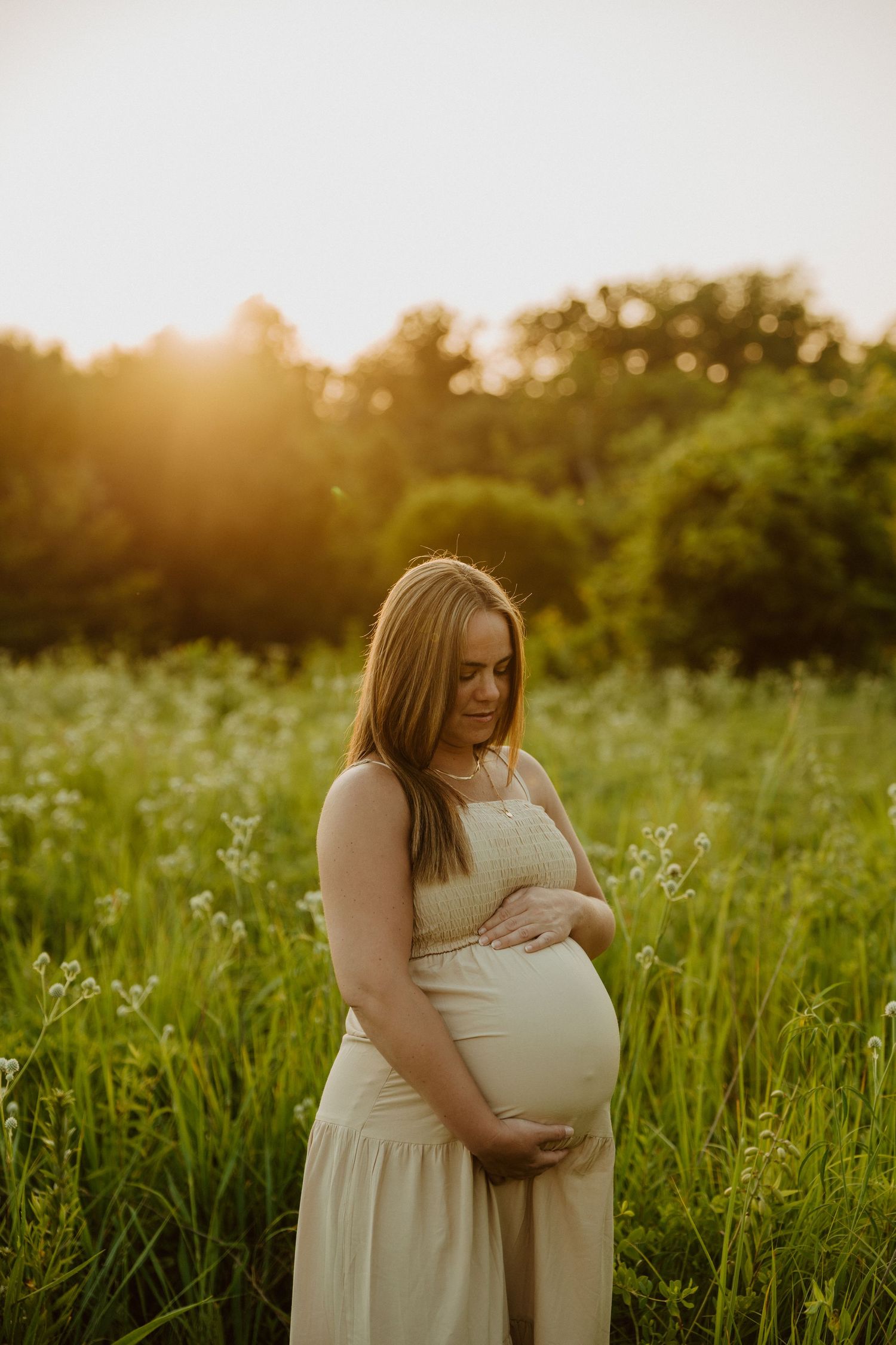 A person in a white dress cradles their pregnant belly while standing in a sunlit meadow at golden hour.