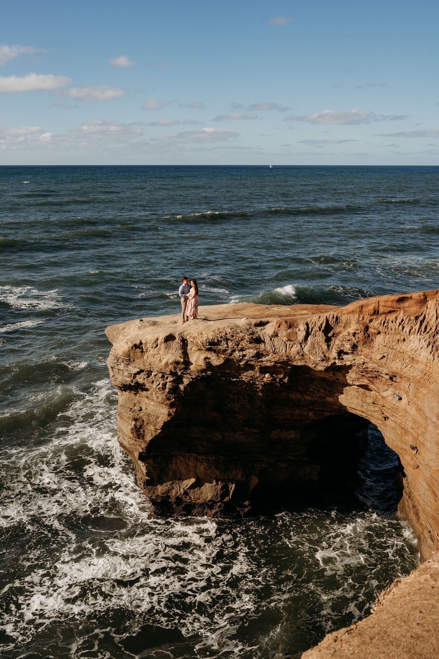 Ocean waves crash against a dramatic rocky arch formation with people standing atop the coastal cliff at sunset.