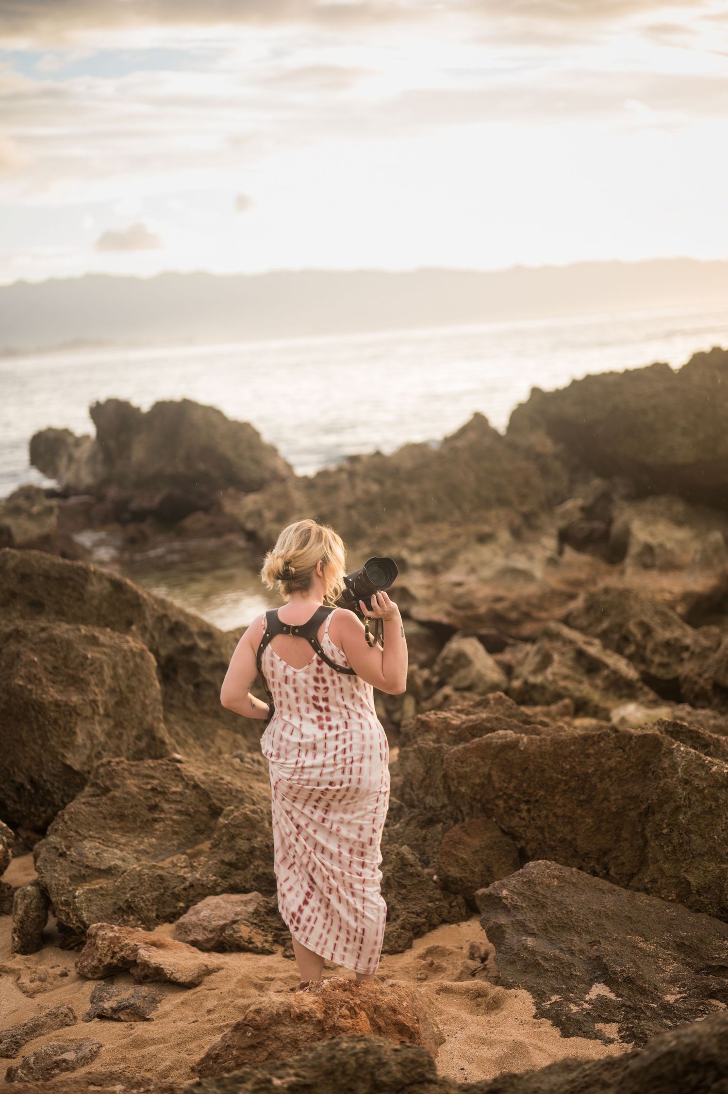 Photographer in flowing polka dot dress stands on rocky coastal terrain during golden hour capturing ocean views.