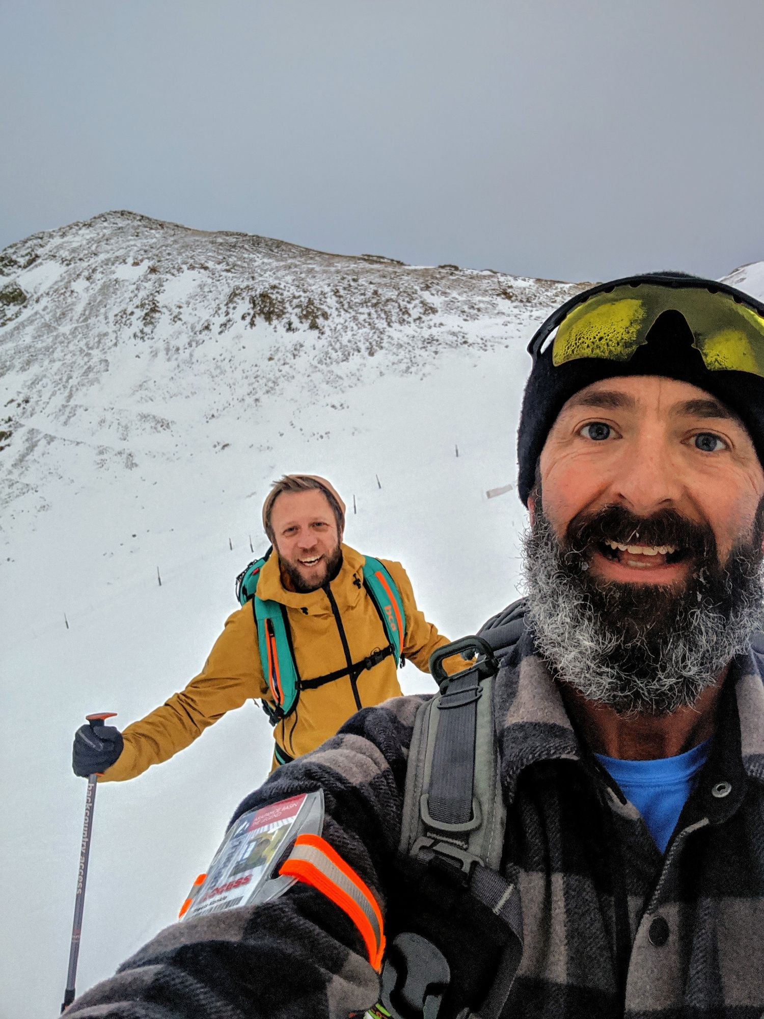 Two winter sports enthusiasts take a selfie while skiing on a snowy mountainside wearing backpacks and ski gear.