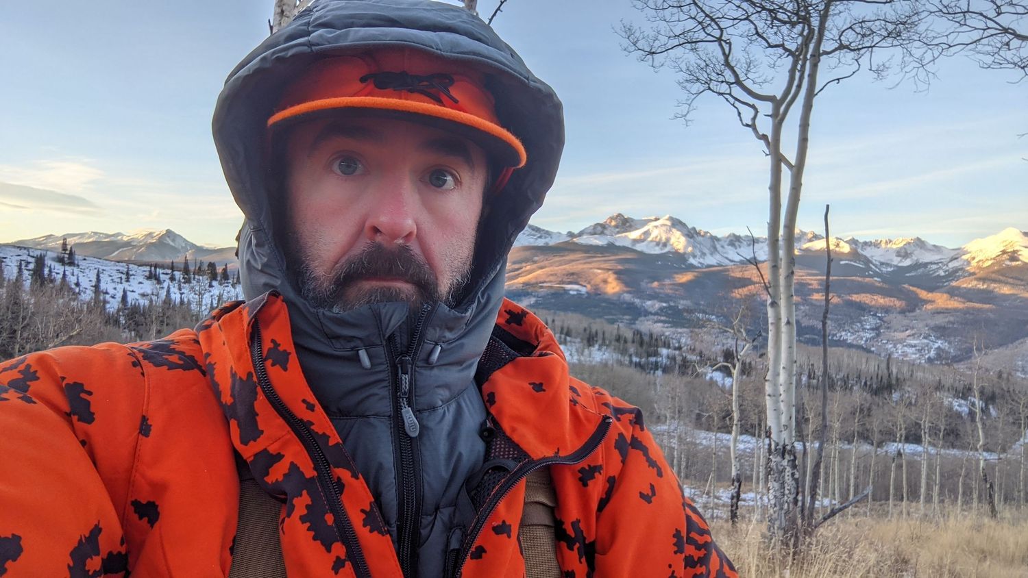 Winter adventurer in orange jacket and black helmet against snowy mountain backdrop during golden hour.